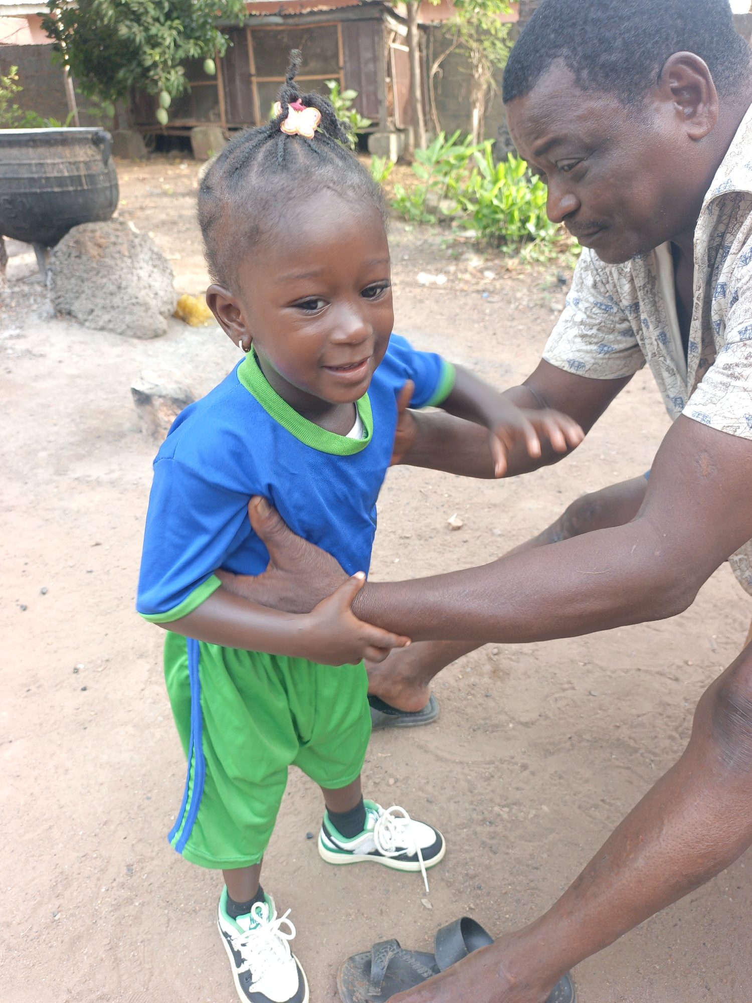 This image, taken in Akwanga, Nigeria, depicts a tender interaction between an adult male and a young child in an outdoor, semi-rural setting.
The main subjects are a man and a child. The man, with dark skin, short dark hair, and a mustache, is wearing a light-colored patterned short-sleeved shirt and dark sandals. He is crouched down, looking gently at the child, and his arms are around her. The child, likely a young girl, has dark skin, braided hair with a colorful butterfly clip, and small hoop earrings. She is dressed in a bright blue t-shirt with green trim, green shorts with a blue stripe, and black, white, and green sneakers. Her face shows a joyful, smiling, or laughing expression as she looks slightly away from the camera.
The activity appears to be a playful or affectionate interaction, possibly with the man guiding or embracing the child. The setting is outdoors on a dirt ground, suggesting a casual, everyday moment. In the background, there are various elements: a large, dark cooking pot resting on rocks, abundant green foliage including a tree bearing green mangoes, and a simple wooden structure or building further back.
Notable details include the bright, natural daylight, indicating a clear day. The rustic backdrop with the cooking pot, dirt ground, and simple structures provides location cues consistent with the provided context of Akwanga, Nigeria. The emotions conveyed are warmth, happiness, and care. There is no visible text in the image.