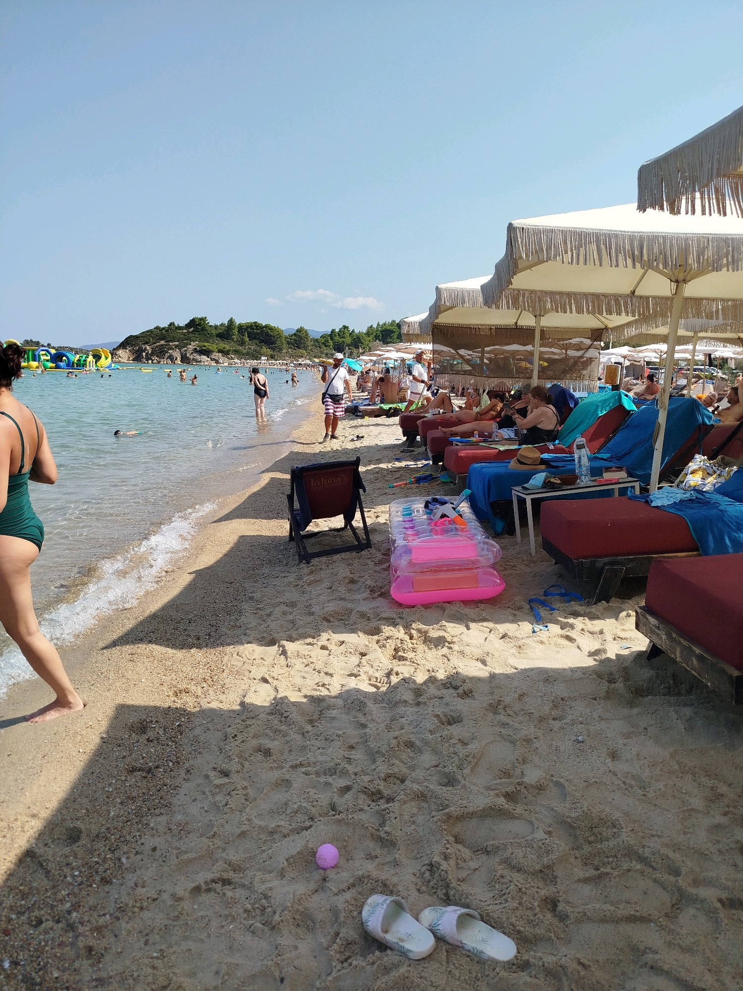 The image captures a sunny day at a beach in the Sithonia Municipal Unit, Greece. The foreground is dominated by sand, with a small pink ball and a pair of flip-flops visible. A woman in a green swimsuit is walking away from the camera towards the water on the left side of the image. The sea is calm with small waves reaching the shore. In the middle ground, several beach chairs and umbrellas are set up, with people relaxing and some walking in the water. Further in the background, there is a rocky island with green trees and inflatable water attractions in the sea. The sky is clear blue.
