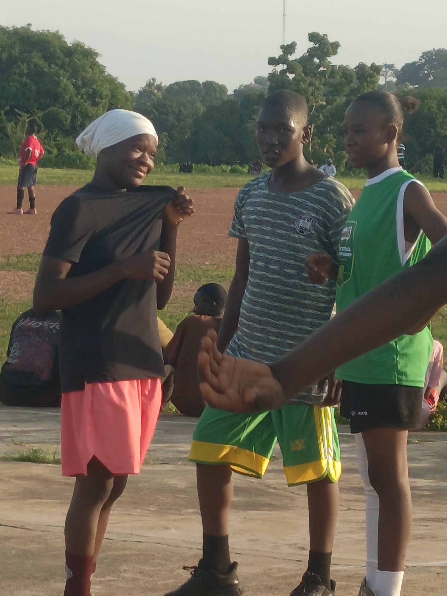 In Akwanga, Nigeria, three young individuals are engaged in an outdoor activity. The primary subjects are positioned in the foreground, appearing to be a group of friends or athletes.
On the left, a person wearing a white headscarf and a black t-shirt is visible, with their left arm extended and hand slightly raised as if in conversation or explaining something. They are also wearing vibrant pink shorts. To their right, a person in a striped t-shirt and green shorts with yellow trim is facing forward, with their hands at their sides. Further to the right, a person in a bright green tank top with white trim is also facing forward. A blurred arm in the foreground suggests another person is present but out of focus.
In the background, a person in a red jersey and dark shorts is standing on a dirt field, with trees and vegetation forming a backdrop under a bright sky. Other individuals are seated or standing further in the background, also appearing to be part of the outdoor activity. The ground surface is a mix of dirt and concrete, indicating an athletic or recreational area. The overall impression is a casual gathering during daylight hours, likely for sports or a similar outdoor event.