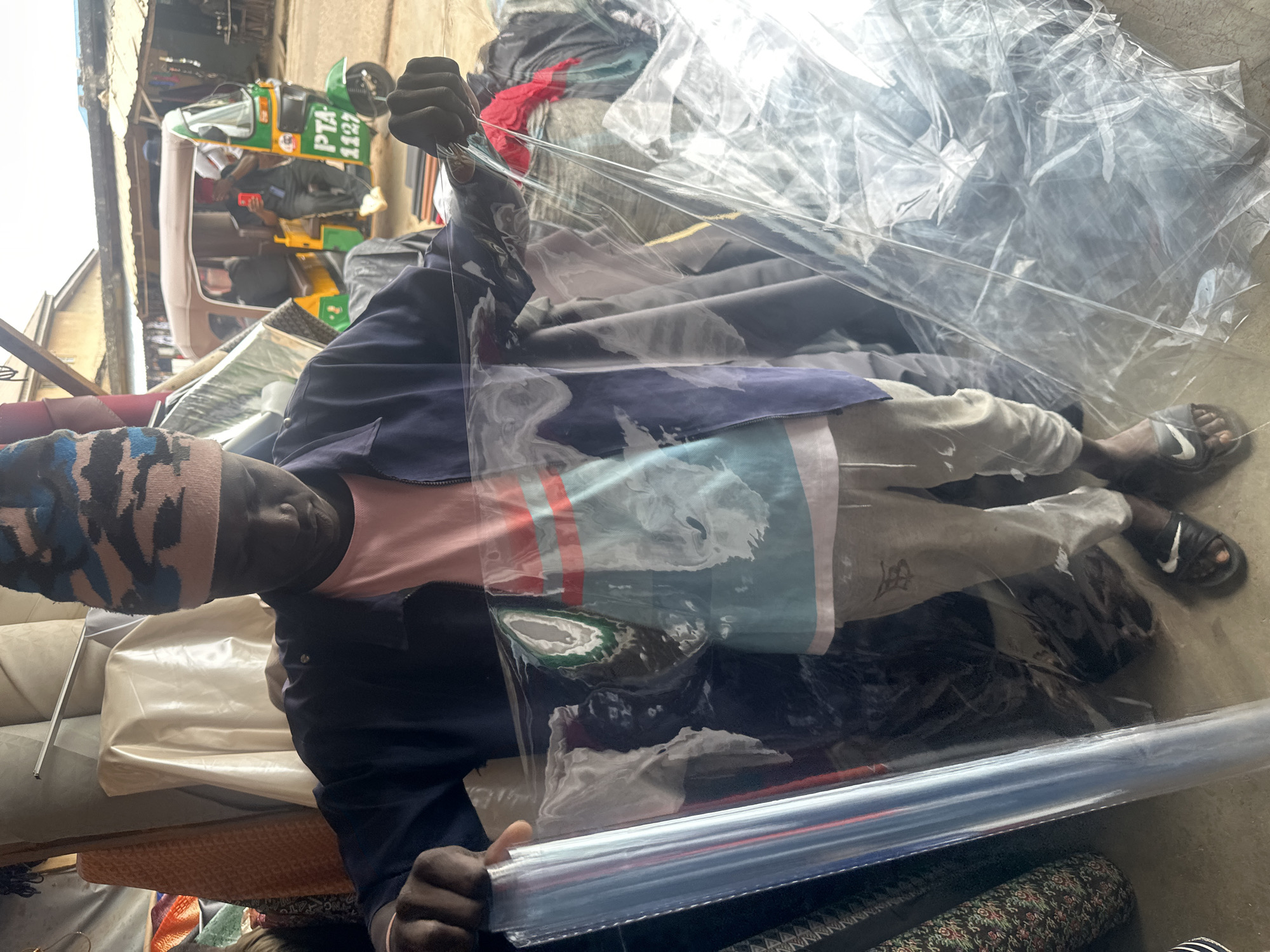 A young, dark-skinned male vendor stands in an open-front market stall in Jos, Nigeria, holding and displaying a large, transparent sheet of plastic film. He wears a camouflage-patterned beanie (blue, pink, black), a dark blue zippered jacket over a light pink and blue striped t-shirt, light grey trousers with a dark graphic on the left thigh, and black flip-flops with a white Nike logo. His left hand holds a tightly rolled section of the plastic, while his right hand holds a more spread-out, crumpled section. Behind him, various textiles and fabrics are stacked in rolls and piles, some patterned, some plain, suggesting an upholstery or fabric shop. Beyond the stall's opening, a bustling street scene unfolds under natural light. A prominent green, yellow, and brown three-wheeled auto-rickshaw, bearing the registration number "PTA 1127," is parked or moving on the unpaved street. Inside the vehicle, a man in a dark shirt is visible, holding a red mobile phone, along with another partial figure. Dilapidated buildings line the background of the street scene.