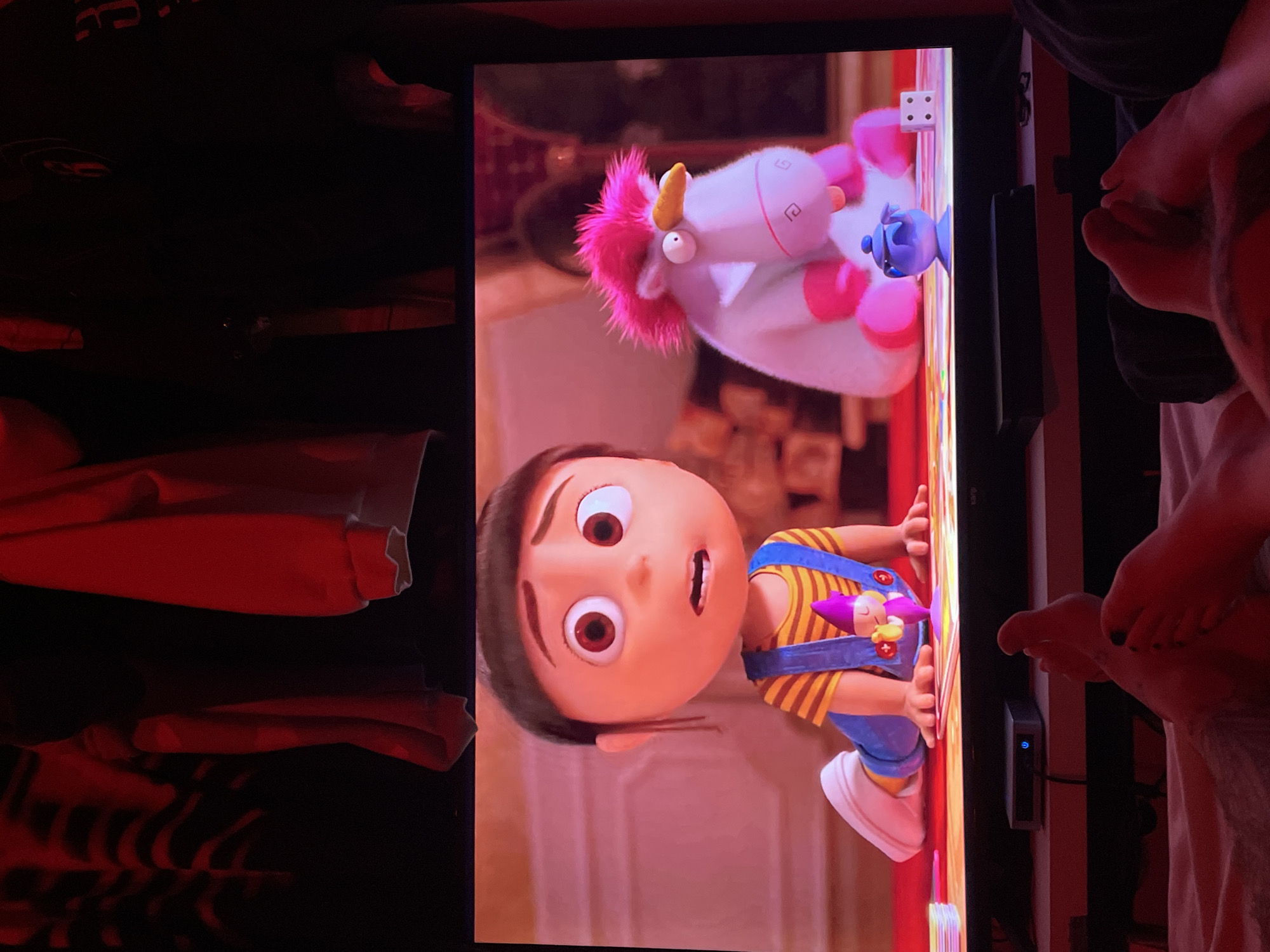 The image shows a television screen displaying a scene from the movie "Despicable Me." The scene features Agnes, a young girl, and a unicorn toy. The television is located in a room with dark, cluttered shelves to the left and a partially visible person's legs and feet to the right. The person's toenails are painted black. There is a device mounted on the back of the TV with a blue power light. The location is not identifiable as being in Buenos Aires, Argentina.