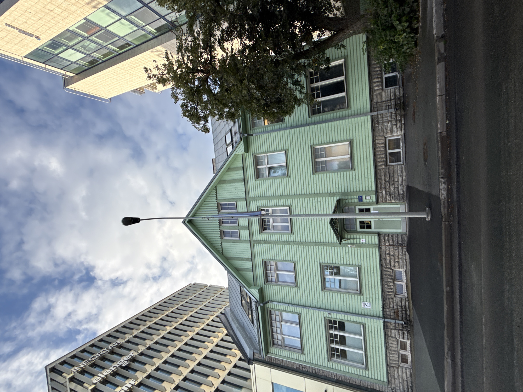 The image depicts a street view in Tallinn, Estonia, featuring a distinctive light green wooden house situated between two modern buildings. The wooden house, with its gabled roof and multiple windows, appears older and contrasts sharply with the contemporary architecture flanking it. The building on the left is a tall, multi-story structure with a facade composed of alternating light-colored panels and dark window sections, displaying the text "INFORMATELENGER" vertically. The building on the right is also a modern, multi-story edifice with a facade primarily of glass windows arranged in a diamond pattern. A mature tree with dense foliage is visible to the right of the wooden house, partially obscuring the modern building behind it. A street lamp stands in front of the wooden house, and the scene is set against a sky with scattered clouds. The foreground shows a paved street and sidewalk.