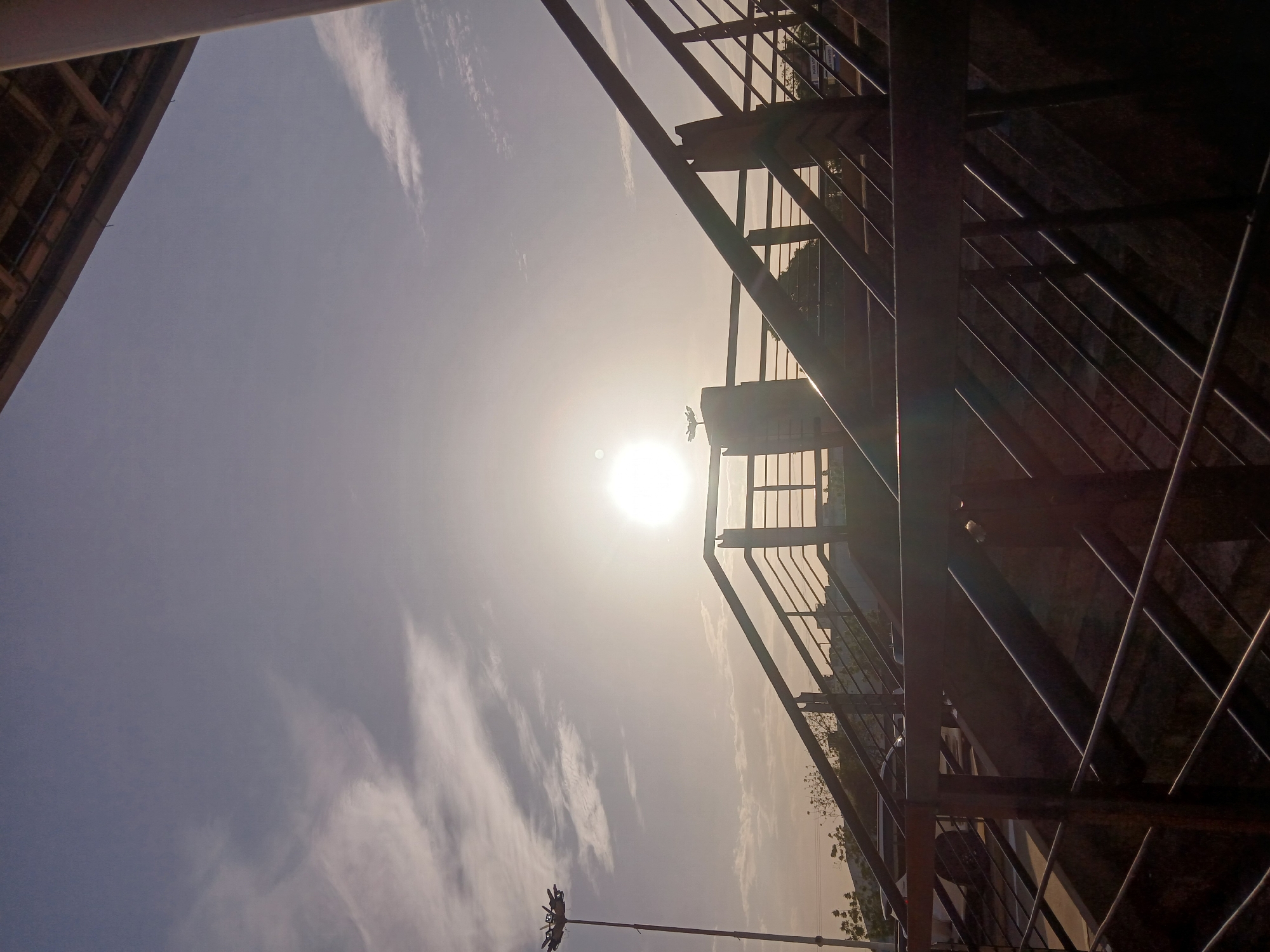 This is an upward-angled, rotated view of an urban scene in Ciudad Guayana, Venezuela. The composition is dominated by a metal structure, likely a staircase or scaffolding, with vertical and diagonal bars casting shadows and creating a complex pattern. The light source, the sun, is visible through the structure, creating a strong glare. The sky is light blue with scattered clouds. Parts of buildings are seen, obscuring the sky. In the bottom of the image, a street light is visible.