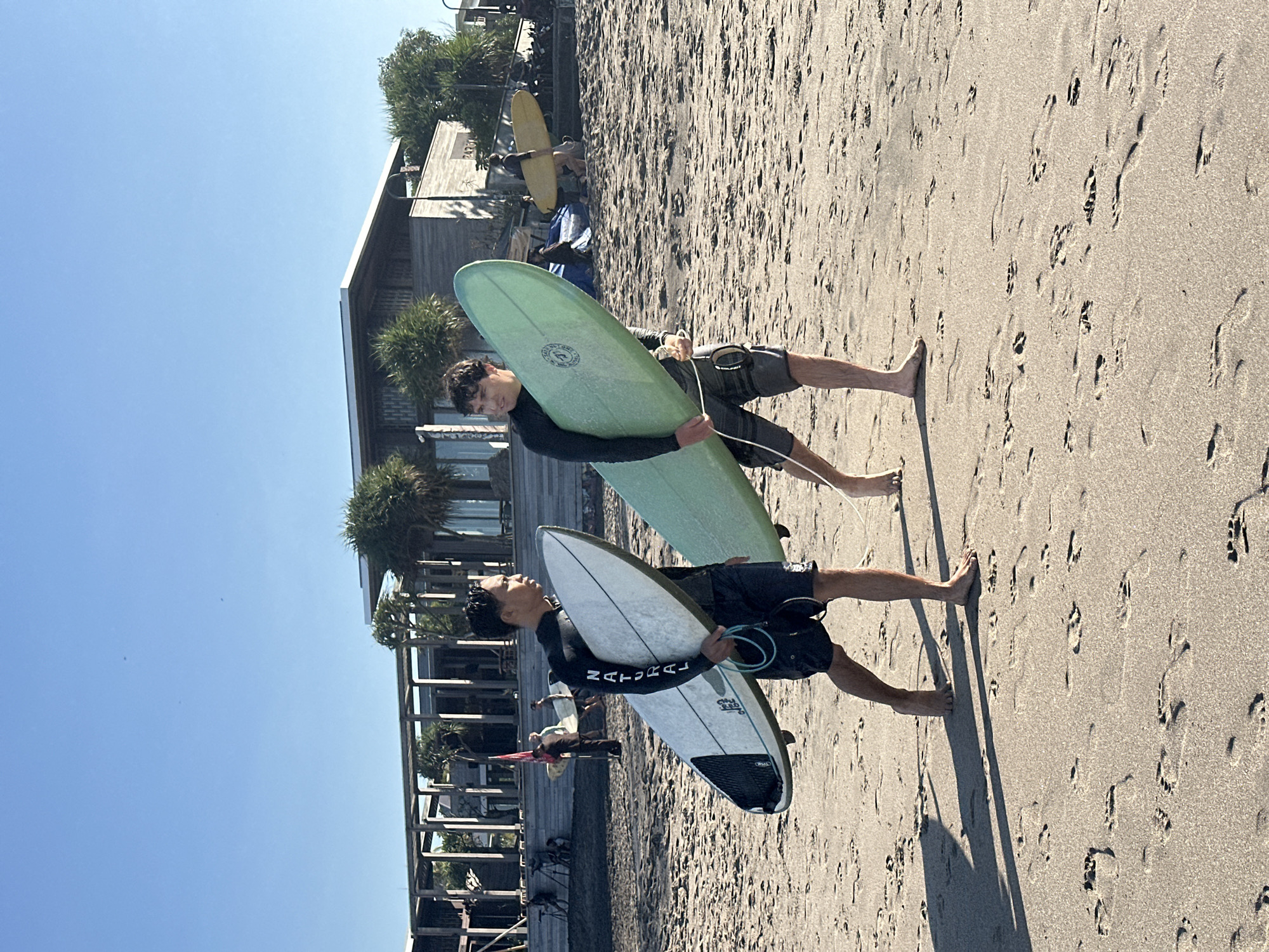 Two young men, presumably surfers, walk across a sandy beach. The man on the left is carrying a white surfboard with black accents and the text "NATURAL" in black lettering down the side. He is wearing a black wetsuit top and dark swim trunks. The man on the right carries a light green surfboard. He is wearing a black wetsuit top and patterned swim trunks. Both men are walking from left to right in the frame. In the background, a modern building with a wooden exterior and a flat roof is visible, surrounded by palm trees. The sky is clear and blue, suggesting a sunny day. The beach is covered in sand with numerous footprints. Other people and surfboards are visible further in the background. The scene appears to be in a tropical location, consistent with North Kuta, Indonesia, a region known for its surf spots.