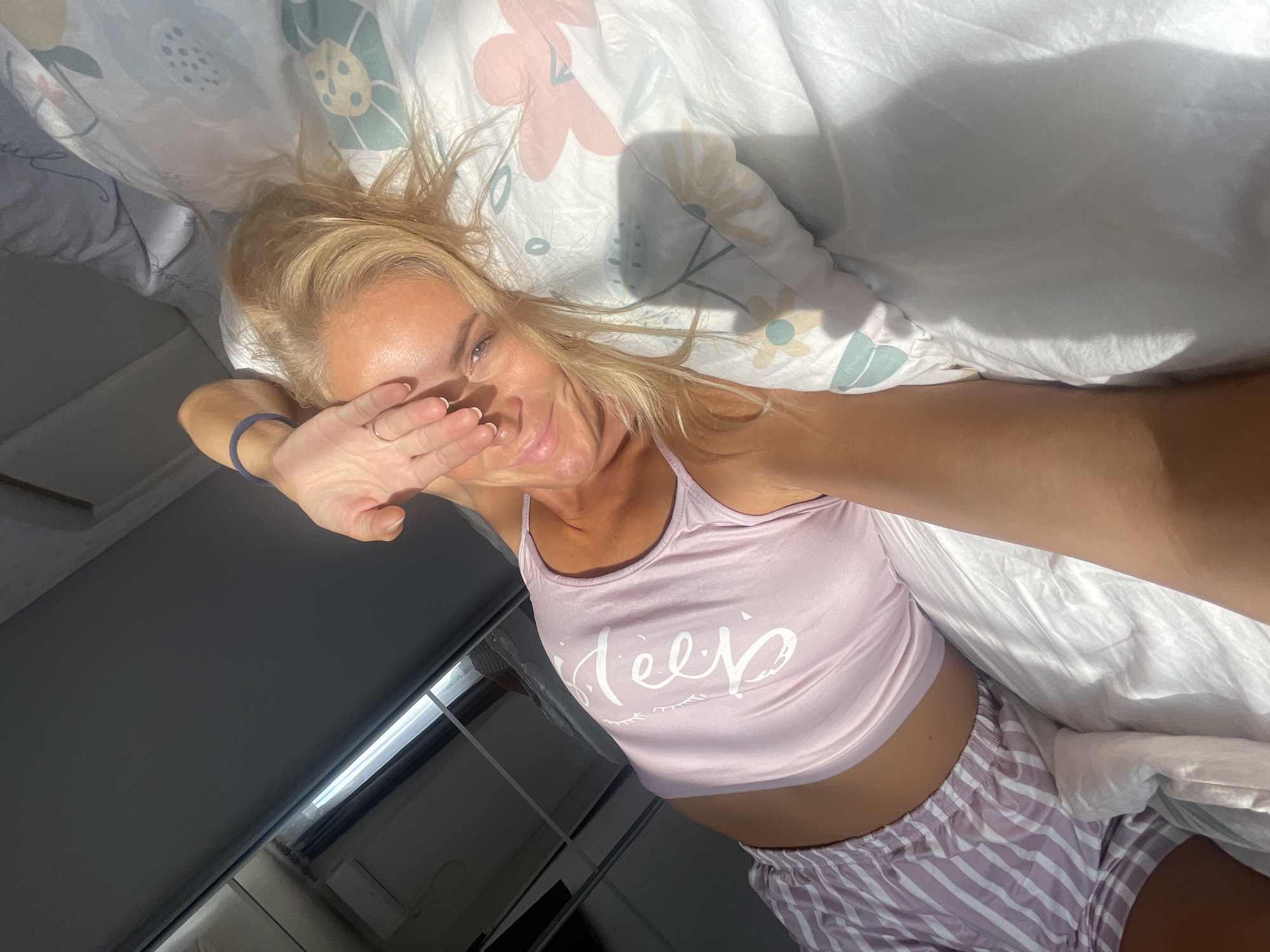 A woman is lying in bed, covering her eyes with her hand. She is wearing a pink tank top with the word "Sleep" printed on it in white, and pink and white striped pajama shorts. The bed has white sheets and a duvet with a floral pattern. Sunlight streams in from the left side of the frame, casting shadows on the wall and bed. The background appears to be a closet with mirrored doors. The overall scene suggests a bedroom in a home. The location is stated to be Baranovka, Russia.