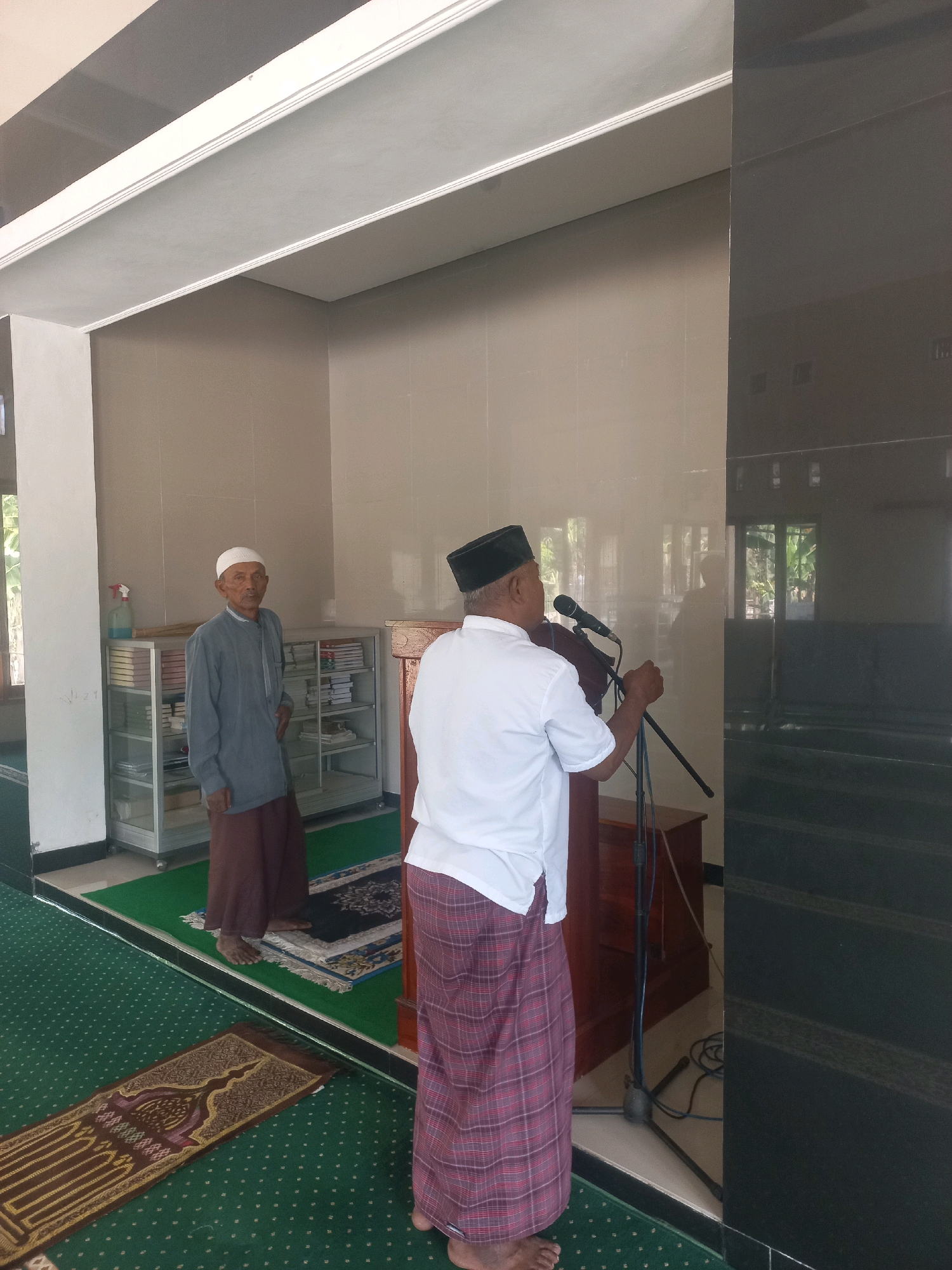 In a room with tiled walls, two men wearing traditional Islamic attire are present. One man, standing to the left, wears a gray long-sleeved shirt and a brown sarong, with a white peci on his head. He stands near a glass display cabinet filled with books. The other man, facing away from the camera, is dressed in a white long-sleeved shirt and a red plaid sarong. He is positioned behind a wooden podium with a microphone on a stand, suggesting he is addressing an audience. The floor is covered with green carpet and prayer mats. The room appears to be a mosque, possibly located in Nguntoronadi, Indonesia, given the context of the question.