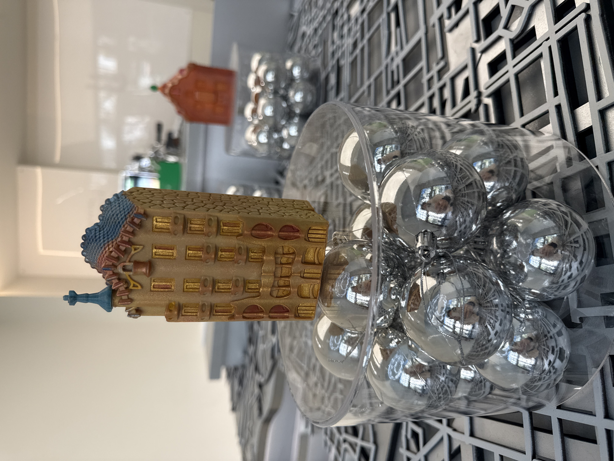 A high-angle, slightly rotated, close-up shot displays three miniature representations of Barcelona landmarks arranged from front to back. The foreground features a model of La Sagrada Familia, colored in brown, gold, and blue, with detailed architectural elements. Adjacent to it is a clear plastic container overflowing with silver baubles, obscuring part of the background. In the back, two more miniature landmarks are seen, one green and one orange, resting on a white ledge or shelf. The background is a grid-like structure, possibly part of the wall, with alternating dark and light squares. The lighting is diffused, creating a soft, reflective shine on the baubles and a distinct contrast between the dark and light areas of the background grid. The architectural models suggest a tourist-oriented or commemorative display, possibly indoors, given the shelf and backdrop.