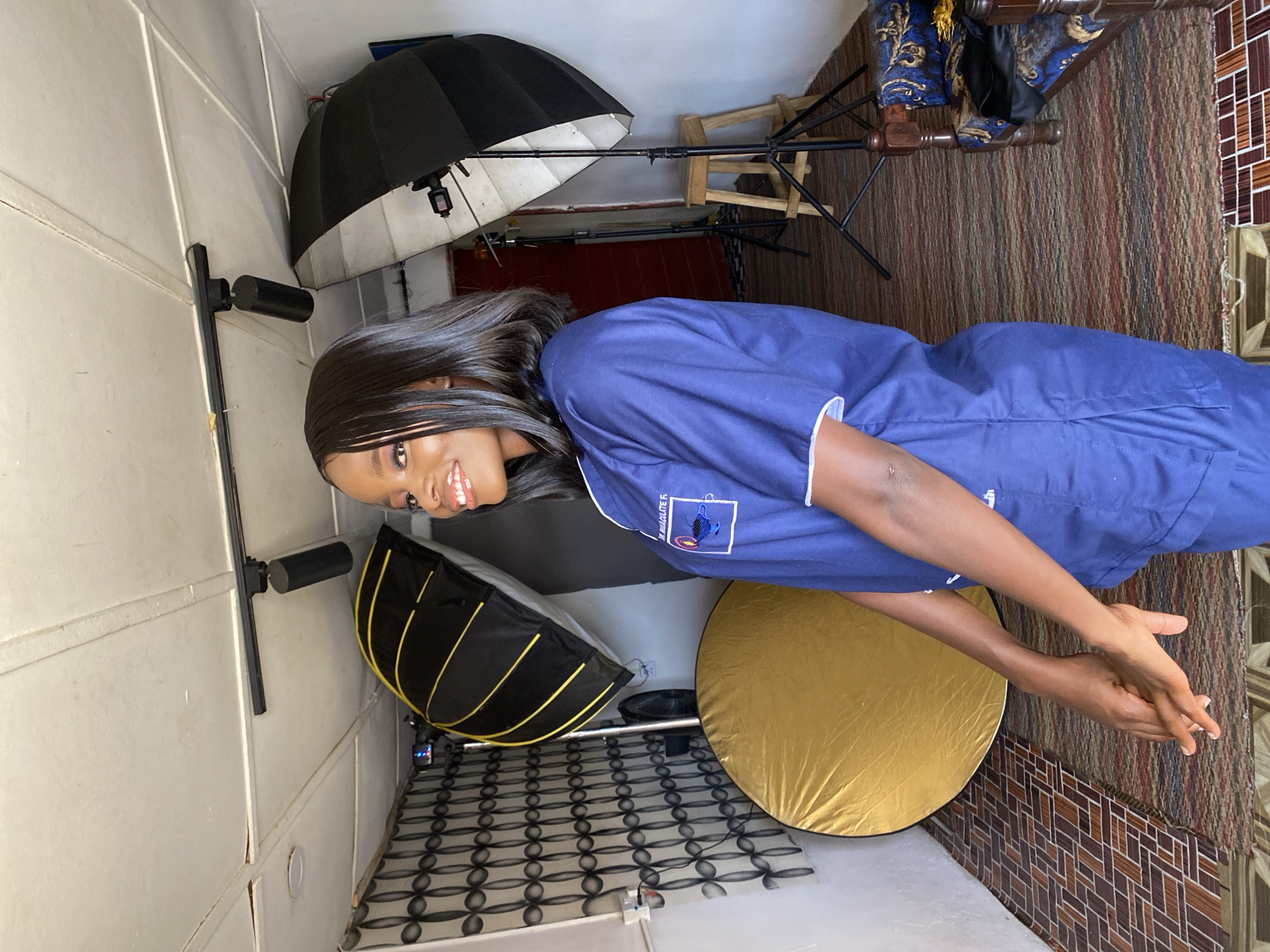 An indoor portrait features a young woman with dark skin and long, straight black hair, smiling warmly towards the camera. She is wearing a dark blue scrub top with white trim on the collar and short sleeves. On her left chest, there is a visible patch with text that appears to read "M IMMACULATE E" and a circular emblem below it, suggesting an institutional or organizational uniform. Her hands are clasped together in front of her.
The setting is a photography studio setup, with various lighting equipment visible around her. To her left, there is a black softbox with yellow accents and a large, round, gold-colored collapsible reflector on the floor. Further back and to her right, two large black studio umbrellas with white inner surfaces are mounted on light stands. One umbrella is more prominent, while the other is partially obscured. Behind the woman, a dark red wall or partition is visible, along with a wooden chair featuring an ornate blue and gold patterned cushion. The floor is covered with a striped brown, red, and black rug, transitioning to a section with a geometric pattern of brown and white tiles or another rug. The surrounding walls are white, made of panels, and show some wear. The overall impression is a posed shot within a functional, if somewhat compact, studio environment.