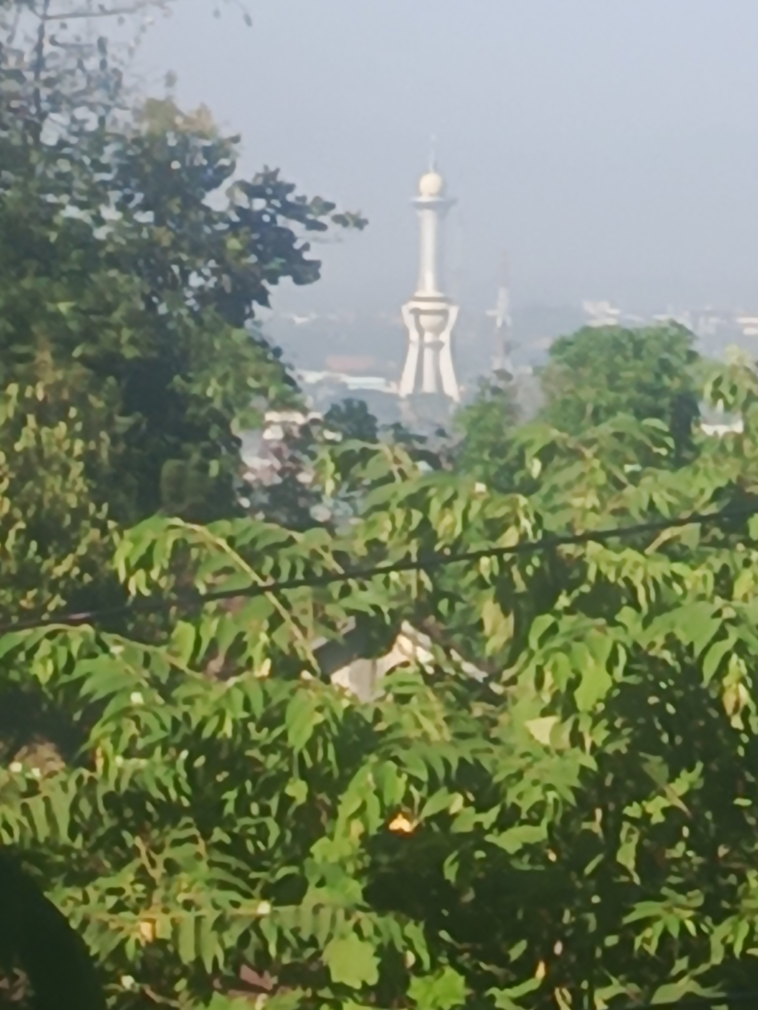 The image is a slightly out-of-focus shot of the Tugu MTQ (Islamic Center Monument) in Kendari, Indonesia, viewed from a distance through lush green foliage. The monument, a tall, white structure with a distinctive dome and ornate base, stands prominently against a hazy sky. The foreground is dominated by the dense leaves of trees and bushes, suggesting a natural vantage point overlooking the city. The overall impression is one of a tranquil, somewhat obscured view of a significant landmark within an urban landscape.