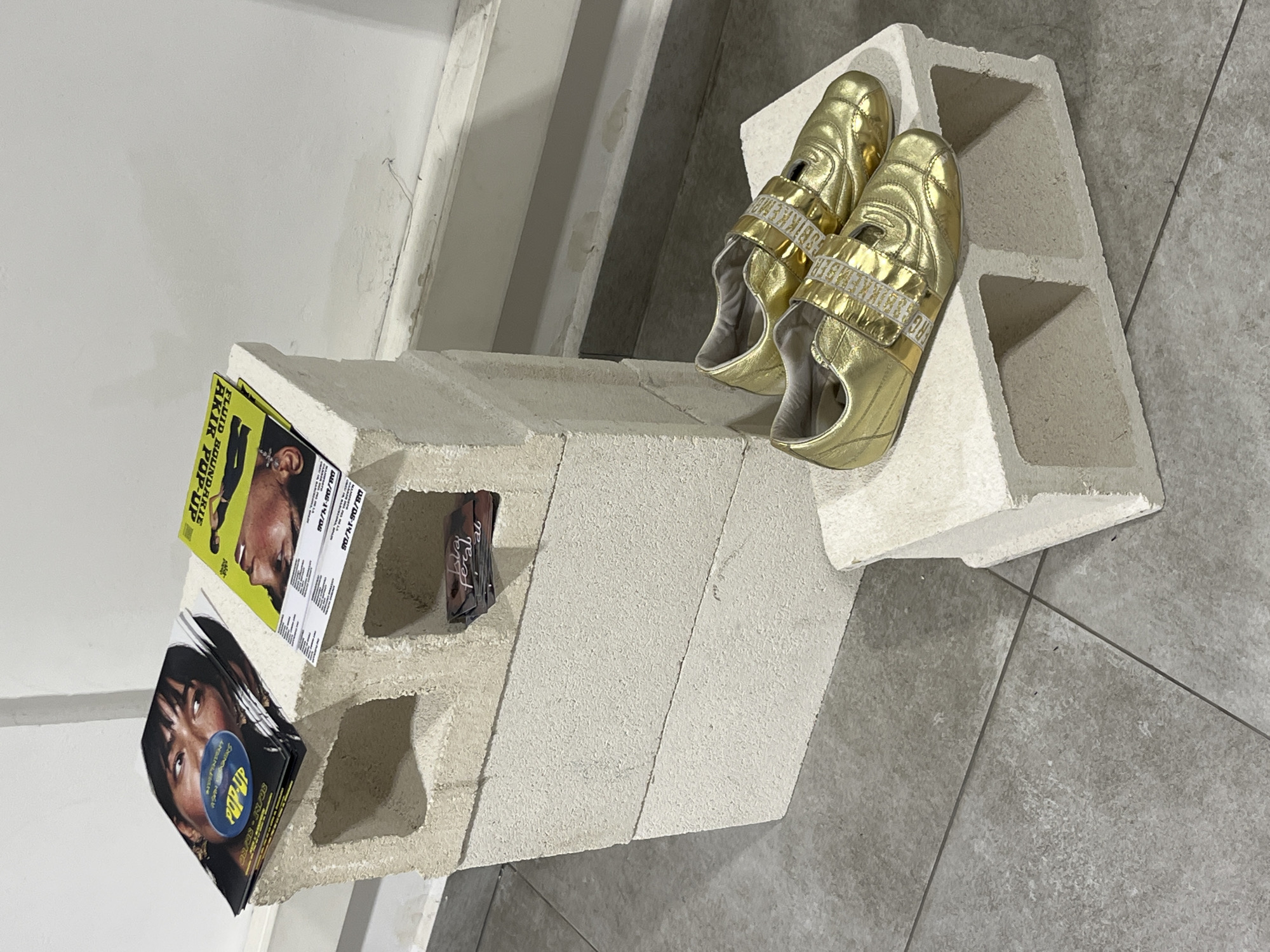 The image shows a still life arrangement indoors. A cinder block, oriented with one of its hollow sections facing up, rests diagonally on the floor. Within the block's hollow sections, various items are arranged: a stack of pamphlets or flyers advertising "Fluid Boundarie Akir Pop-Up," a small card or booklet labeled "bia feral," and a pair of gold-colored shoes positioned on top of the block. The background consists of a gray tiled floor, a white wall, and what appears to be a portion of a staircase or ramp. The image offers no information to determine that the location is Barcelona, Spain.