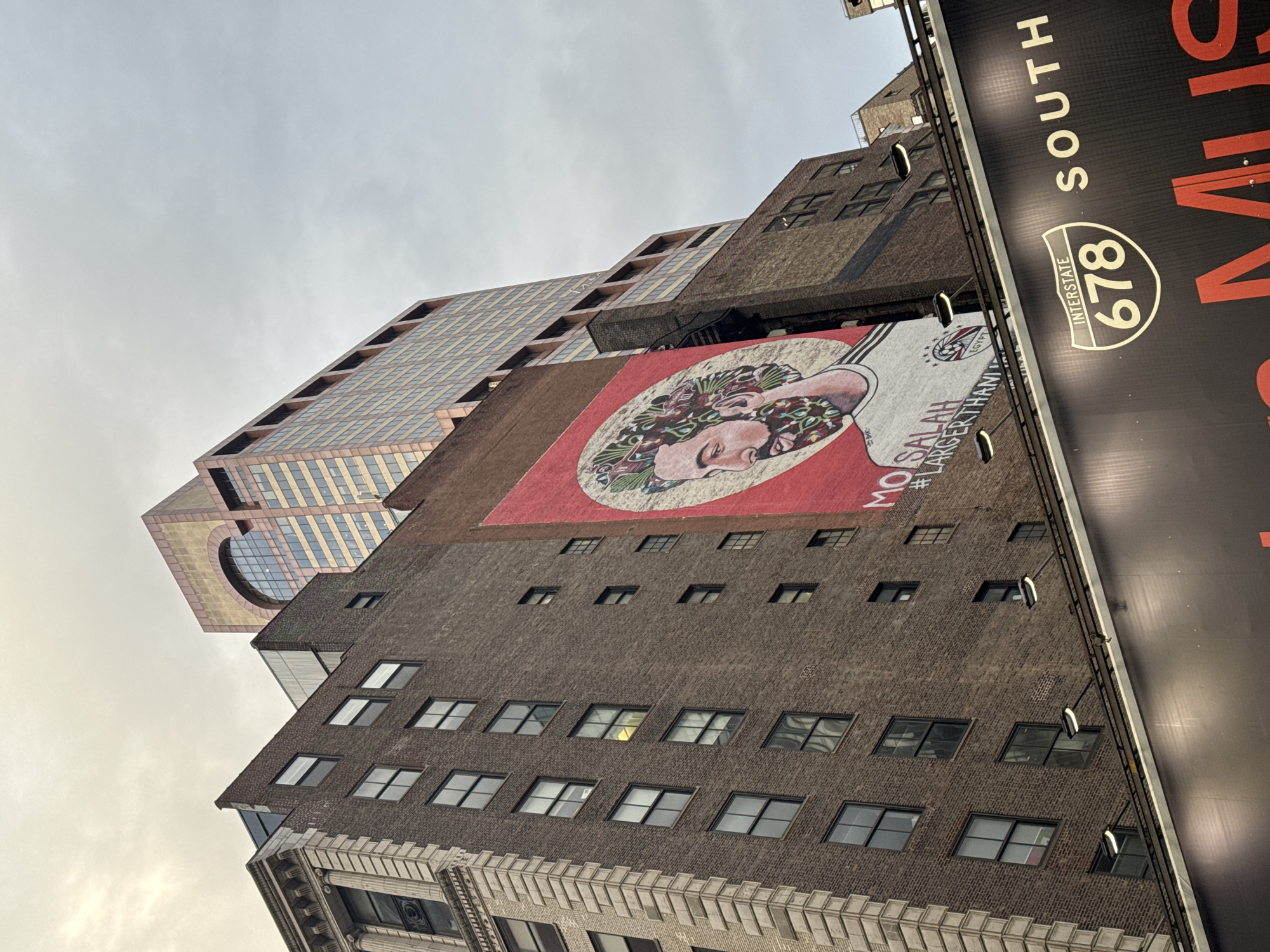 The image displays an upward view of an urban environment in Queens, New York City, United States of America, under an overcast sky.
A prominent mural dominates the mid-ground, affixed to the side of a multi-story brown brick building with numerous rectangular windows. The mural features a stylized portrait of a bearded man, identified by text as "MO SALAH" with the hashtag "#LARGERTHANLIFE," rendered on a red circular background. The man's hair and beard are depicted with intricate, colorful floral and leafy patterns. He wears a light-colored, striped jersey with a small Egyptian flag emblem. To the left, an adjacent brick building exhibits ornate, light-colored stone architectural details including columns and cornices.
In the background, a taller, modern skyscraper with a reddish-brown and beige facade and multiple horizontal windows is visible. Its upper section features a large arched window and a lighter, possibly copper-toned, crown.
At the bottom of the frame, a large, dark billboard is partially visible. It prominently displays the white text "INTERSTATE 678 SOUTH" within a standard interstate shield logo. Several illumination fixtures are mounted on a bar above this billboard, projecting light downwards. The general impression is of a daytime scene under cloudy conditions.