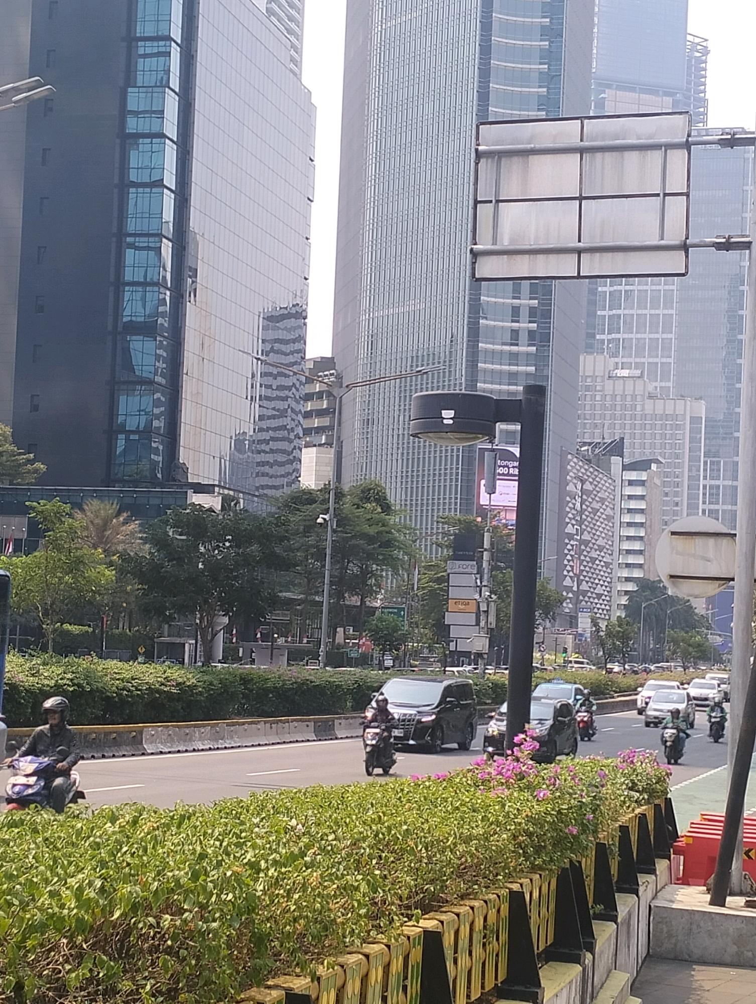 The image captures a bustling street scene in Central Jakarta, Indonesia, characterized by modern high-rise buildings and a busy flow of traffic. A motorcyclist wearing a helmet is seen on the left, navigating the road. Several other motorcycles and cars are also visible, indicating a typical urban commute. Lush green bushes and flowering plants line the road, creating a visually appealing contrast with the concrete jungle of the city. The architecture of the buildings suggests a commercial or business district, with their sleek glass facades reflecting the daylight. The presence of streetlights and signage further emphasizes the urban environment. The overall impression is one of a dynamic and active cityscape.