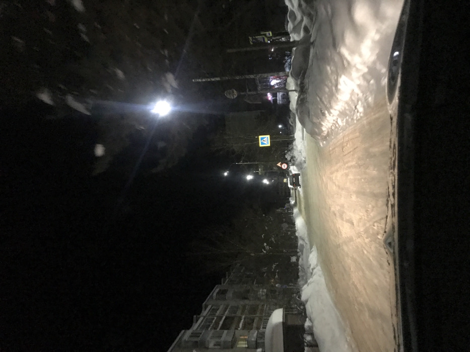 A dark street scene, likely at night, shows a road covered in a slushy, icy mix, with piled snow to the side. A streetlamp illuminates the road. A building stands to the left. Various traffic signs are visible along the road, including a crosswalk sign. A vehicle appears to be parked on the right side of the street. The image is taken from inside a car, with part of the car’s interior visible on the right.