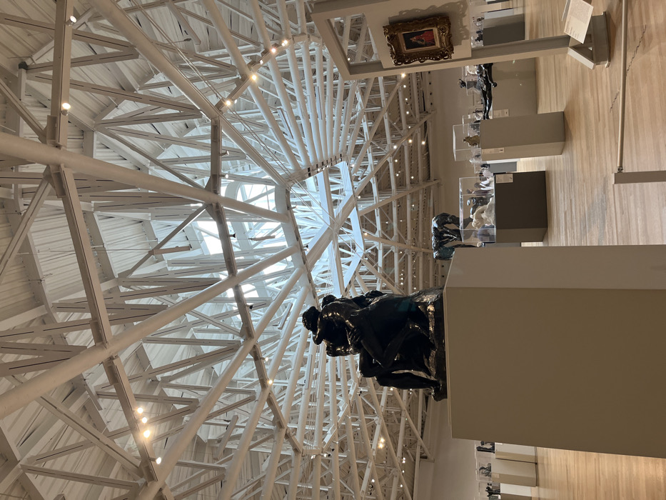The museum's interior features a striking white geometric ceiling, contrasting with the artwork displayed below. A dark sculpture prominently sits atop a light-colored pedestal. Other artworks, enclosed in glass cases, are strategically placed along the wall, while a painting hangs higher up. The wooden floor reflects the ambient light. The ceiling structure and artwork create a visually dynamic and engaging space.