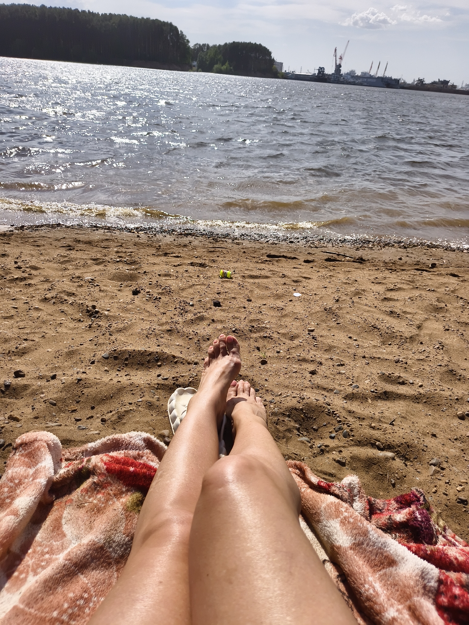 A first-person perspective shot captures a scene on a sandy beach with a body of water and industrial elements in the background. A person is lying on a blanket with their legs visible in the foreground. The blanket is multi-colored, featuring shades of beige, white, and red. The person's feet are bare, and a white sandal is visible near their legs.
The beach is composed of tan sand with small pebbles and debris scattered across it. In the distance, a body of water stretches to the horizon, reflecting sunlight. Beyond the water, there is a line of trees and industrial infrastructure, including cranes, suggesting a port or industrial area. The sky is partly cloudy.