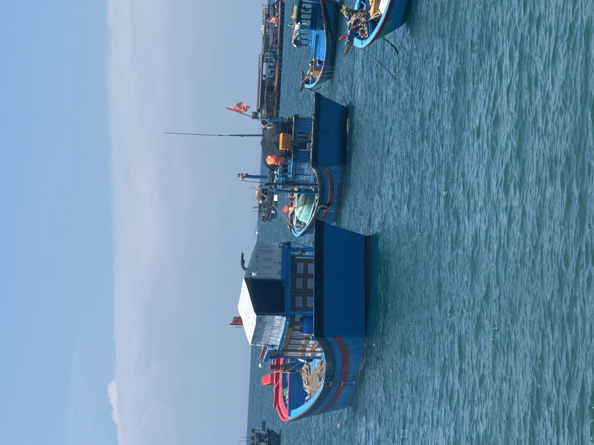 The image depicts a fleet of blue fishing boats anchored in clear blue ocean water under a bright, partly cloudy sky. The boats are of various sizes and designs, with some featuring red trim and white roofs. One prominent boat in the foreground is a medium-sized vessel with a blue hull, red stripes, and a white, box-like structure on its deck. It has a ladder extending from its side into the water and appears to be carrying fishing gear. Another boat to its right is smaller and also painted blue. In the background, larger, more industrial-looking barges are visible, suggesting a working harbor or anchorage. Red flags are flying from some of the boats. The water is choppy with small waves and reflections of the sky and boats. The scene conveys a sense of a working fishing community in a coastal area.