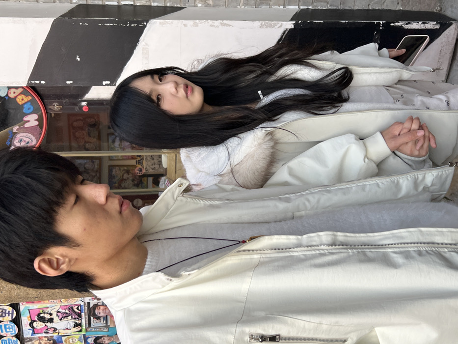 Two individuals, an Asian male and an Asian female, are depicted standing outdoors in an urban environment in Yuzhong District, China. The male, positioned on the left, has dark hair and wears a light-colored zip-up jacket over a light-grey sweater, with a dark cord necklace displaying a light-colored pendant. He is observed looking downwards and to his right. The female, positioned on the right, has long dark hair and wears a matching light-colored hooded jacket featuring a light brown fur-lined hood, over a lighter-toned top and patterned light-colored pants. She holds a dark-colored smartphone in her right hand and is looking to her right with a neutral expression. Their left hands are interlocked. The background behind the male consists of a storefront window displaying various colorful illustrations, possibly anime or cartoon art, along with partially visible Chinese character signage. Behind the female is a wall featuring a prominent black and white geometric pattern.