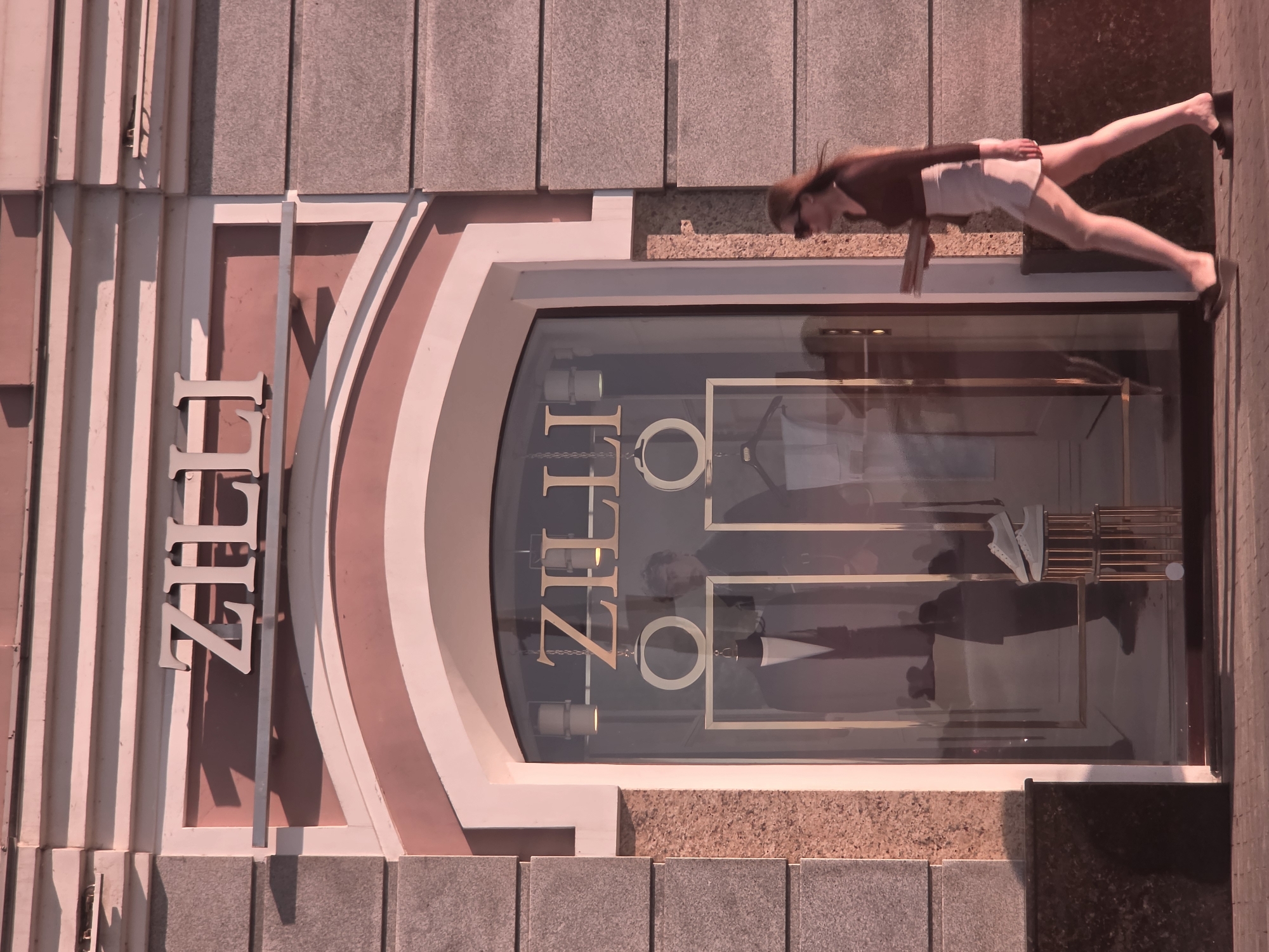 A woman walks past a store window with the name "ZILLI" displayed above and within. The store appears to sell men's formal wear, as evidenced by the mannequins dressed in suits visible through the window, along with a pair of white sneakers displayed on a metallic stand. The building's facade is constructed with large, textured stone blocks and features an arched entryway. The lighting suggests it is daytime, possibly late afternoon, with long shadows cast across the pavement. The woman is wearing a dark long-sleeved shirt, a light-colored skirt, sunglasses, and dark wedge sandals. She is carrying what appear to be several flat objects, possibly books or a tablet, in her left hand. The scene is situated on a city street.