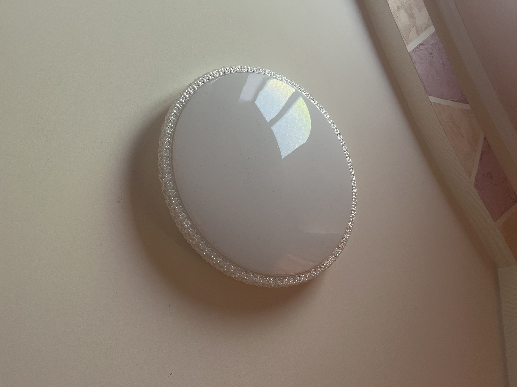 The image shows a close-up, overhead view of a flush-mount ceiling light fixture. The fixture has a white, opaque diffuser with a decorative trim of clear, faceted embellishments around its perimeter. The ceiling is painted a light, neutral color, with subtle shadows cast by the light fixture. In the lower right quadrant of the image, a portion of a decorative border or valance is visible, featuring a textured pattern with shades of pink and beige. The scene appears to be an interior, likely a residential setting. No people or actions are depicted. The location is identified as Tarnivtsi, Ukraine.
