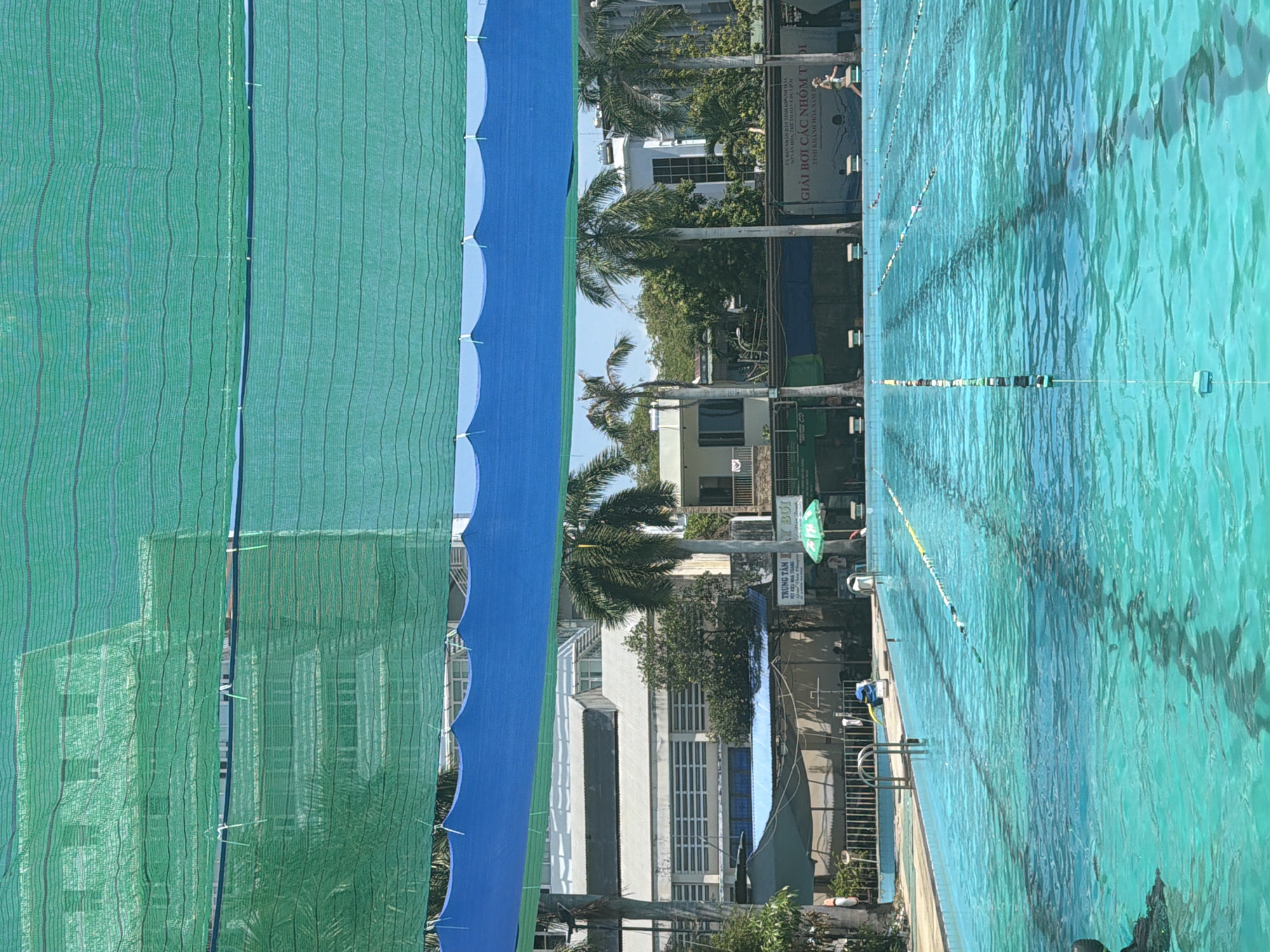 The image shows an outdoor swimming pool in Nha Trang Ward, Vietnam. The pool is divided into lanes by buoys and lane ropes. The water is turquoise and appears calm with ripples. In the background, there are several buildings, palm trees, and lush green vegetation. Two large green and blue nets are stretched across the top of the image, providing shade over the pool. A sign with Vietnamese text is visible on the right side of the image, with the words "GIẢI BƠI CÁC NHÓM TUỔI" which translates to "Giải bơi các nhóm tuổi" or "Age Group Swimming Competition". A woman is seen walking along the edge of the pool on the right side. There are several swim lane markers visible in the pool. The overall scene suggests a recreational or training facility.