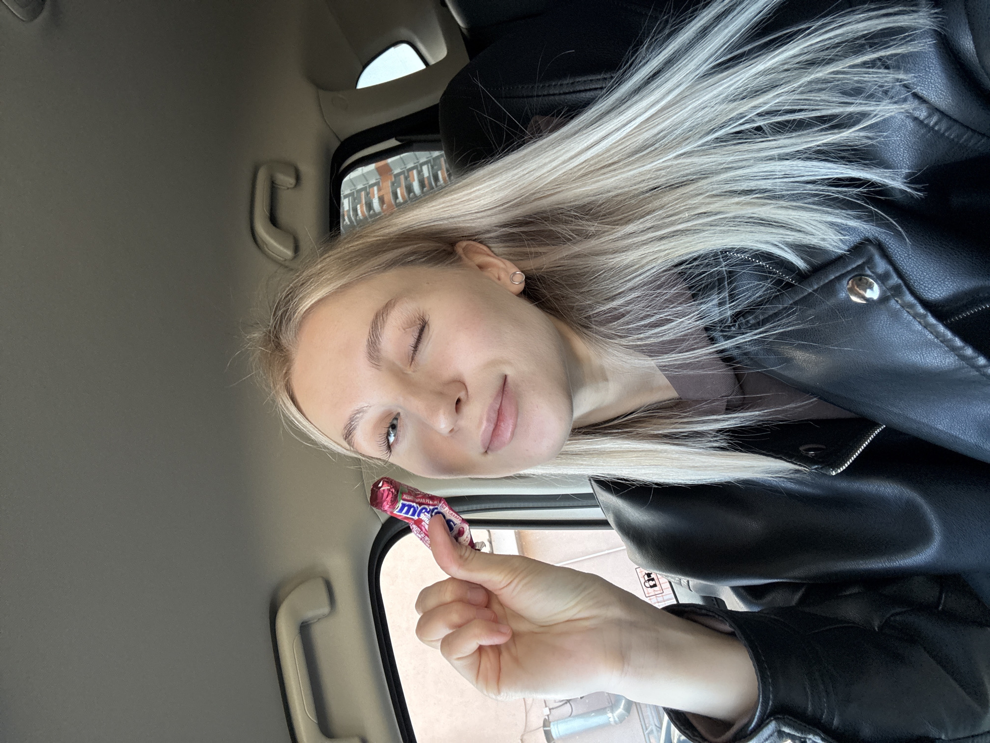A young woman with blonde hair and a black leather jacket is sitting in a car, possibly in Izhevsk, Russia. She is holding a 'merci' chocolate candy bar in her right hand, and winking with her left eye. The interior of the car is visible, including the seat, window, and ceiling.