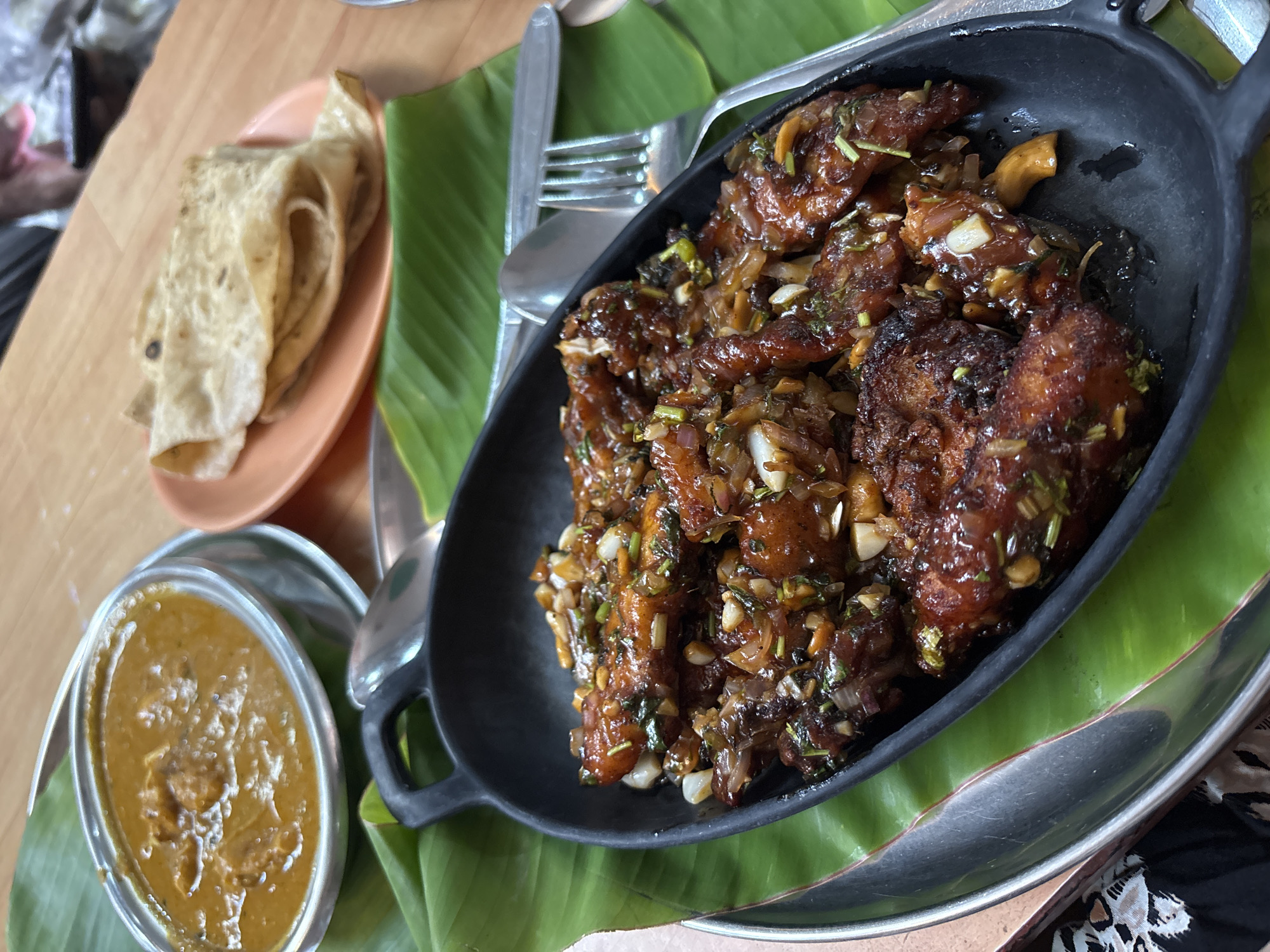 The image shows a table setting with Indian cuisine. A black oblong dish containing what appears to be chilli chicken sits on a large green leaf, likely a banana leaf, placed on a silver tray. A fork and spoon rest diagonally across the dish. To the left of the black dish, there is a small, silver dish containing a curry or lentil soup. Further to the left and slightly behind, a stack of flatbread, possibly roti or chapati, is placed on a peach-colored plate. The food is presented on a wooden table. The setting is likely in a restaurant, and the location is given as Kamalapura, India, although this is not directly verifiable from the image itself. The scene suggests a meal is being served, with different components of a traditional Indian meal arranged together.