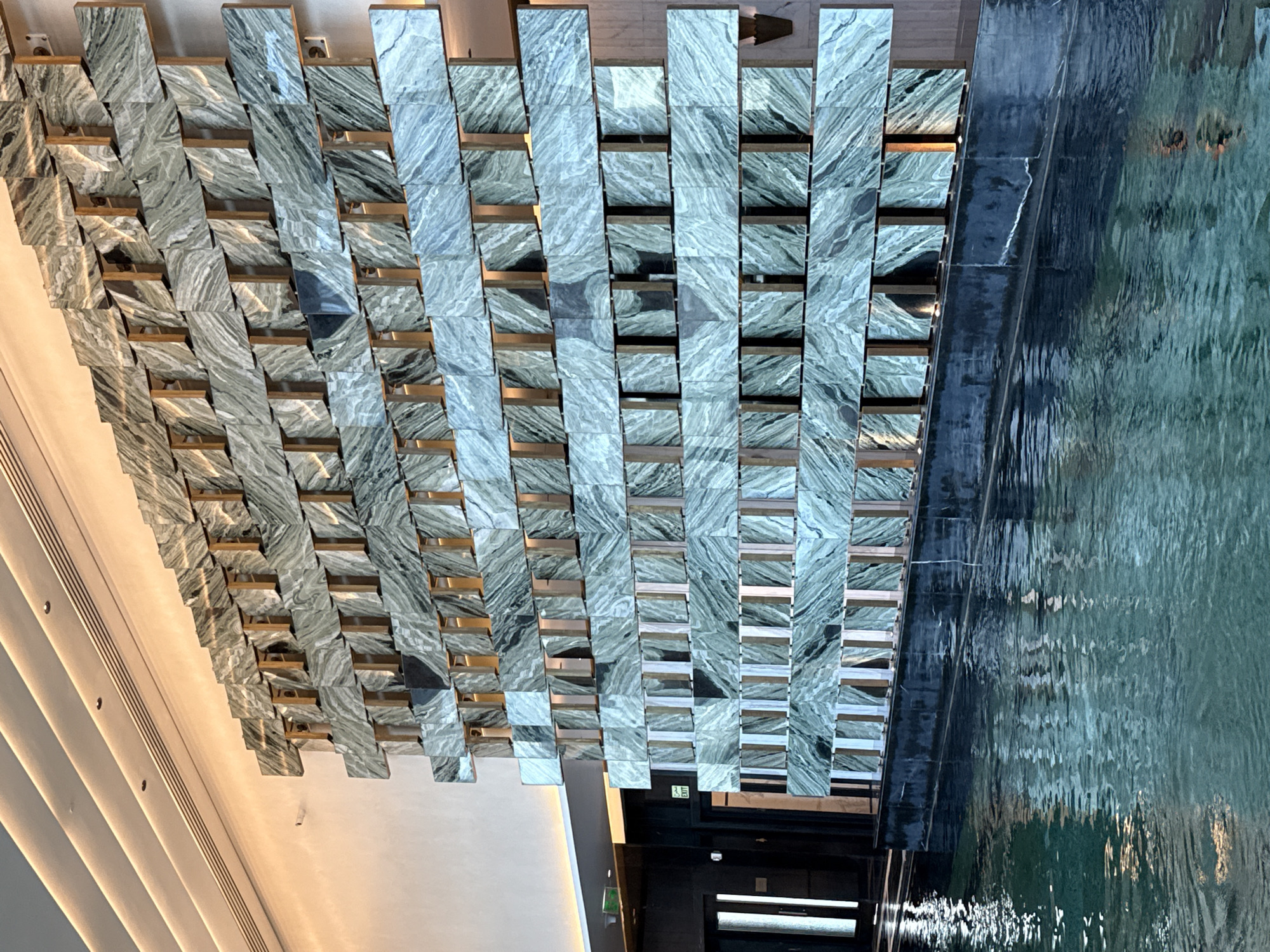 A low-angle shot depicts a decorative wall made up of stacked, rectangular, green-and-gray marble tiles separated by gold-toned strips, resembling a woven pattern. The wall is adjacent to a flowing water feature, reflecting the surrounding environment and obscuring any details behind it. Below the decorative wall is a dark doorway with a green emergency exit sign above. The overall location appears to be an interior space in a modern building. The specific city location is unknown.