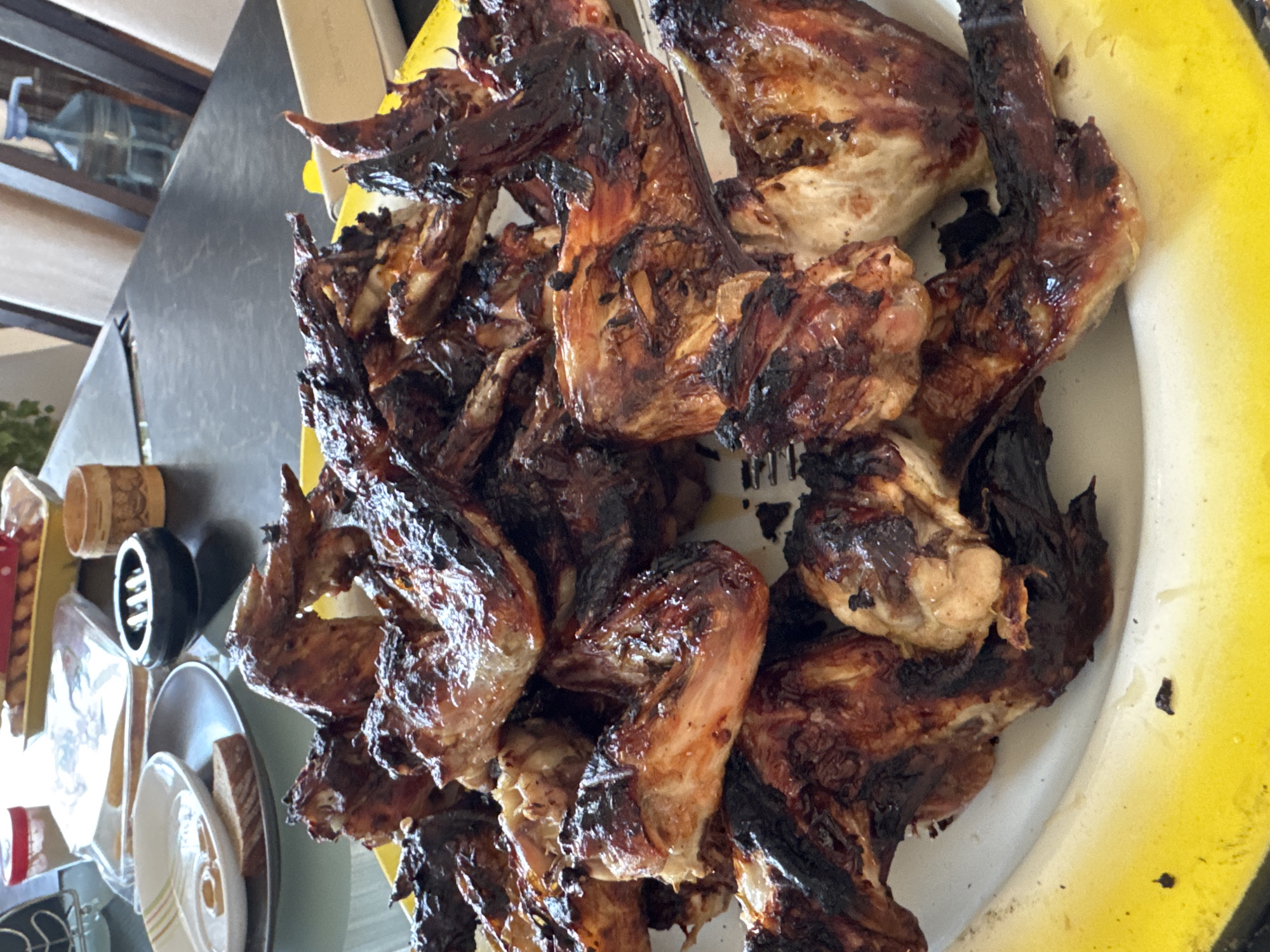 A high-angle, close-up shot depicts a large serving platter piled high with what appears to be barbecued chicken wings. The wings are darkly browned, appearing somewhat charred in places. The platter has a white base with a thick yellow rim, and is set upon a dark, possibly metallic surface. To the left of the platter are plates and a cylindrical object. The location cannot be determined from the image.