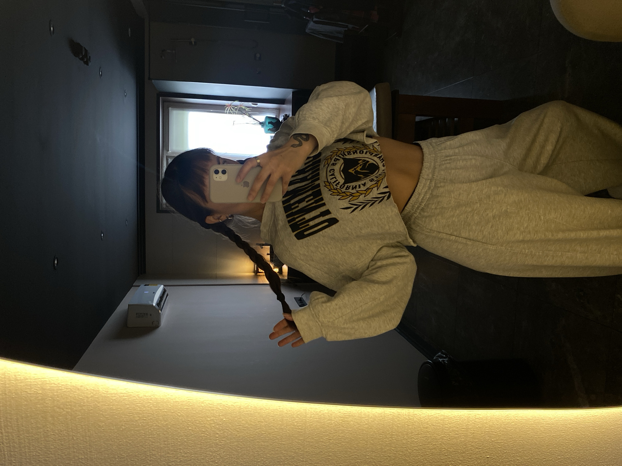 In a dimly lit room, a young woman stands facing a mirror, taking a selfie. She has dark hair styled in two braids and is wearing a light gray cropped sweatshirt with a circular graphic and matching sweatpants. Her left hand is raised, touching one of her braids. The room features a large, brightly lit vertical fixture on the left, a wall-mounted air conditioner on the back wall, and a window with blinds in the background. The overall setting appears to be a private residence. No specific details can confirm the location within Votkinsk, Russia.