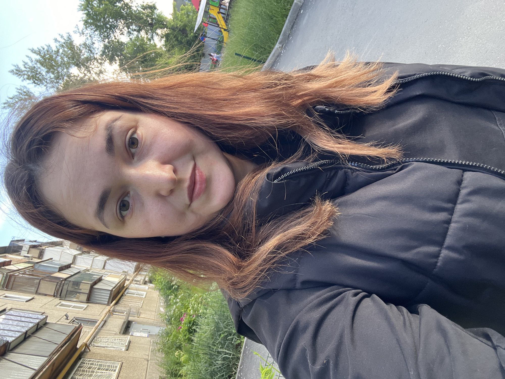A woman with shoulder-length reddish-brown hair is taking a selfie outdoors. She is wearing a black puffer jacket over a black top. In the background, there is a multi-story apartment building with balconies, some of which are enclosed. There is also greenery, a paved area, and a children's playground with yellow slides and a red structure. The lighting suggests it is daytime, possibly late afternoon, given the shadows. The environment appears to be a residential area in Chelyabinsk, Russia, characterized by Soviet-era apartment blocks and communal outdoor spaces.