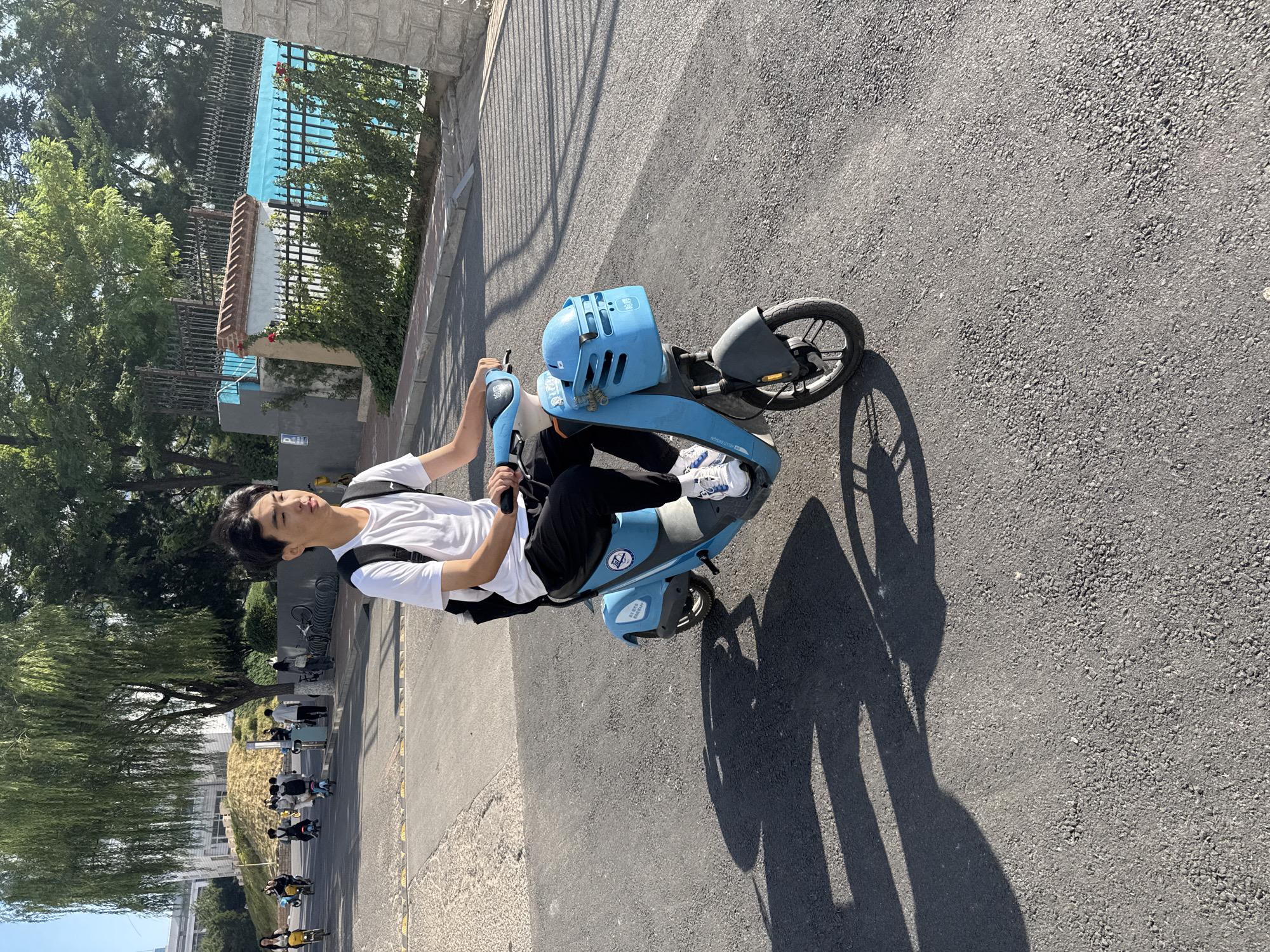 An East Asian male is riding a blue electric scooter on an asphalt road. He is wearing a white t-shirt, black pants, and white sneakers, and has a black backpack on. The scooter has a basket in the front with a blue helmet inside. In the background, there are trees, buildings, a fence, and other people on bikes and scooters. The scene appears to be in an urban or campus environment in Zhongtun, China. The sun is casting shadows on the road.