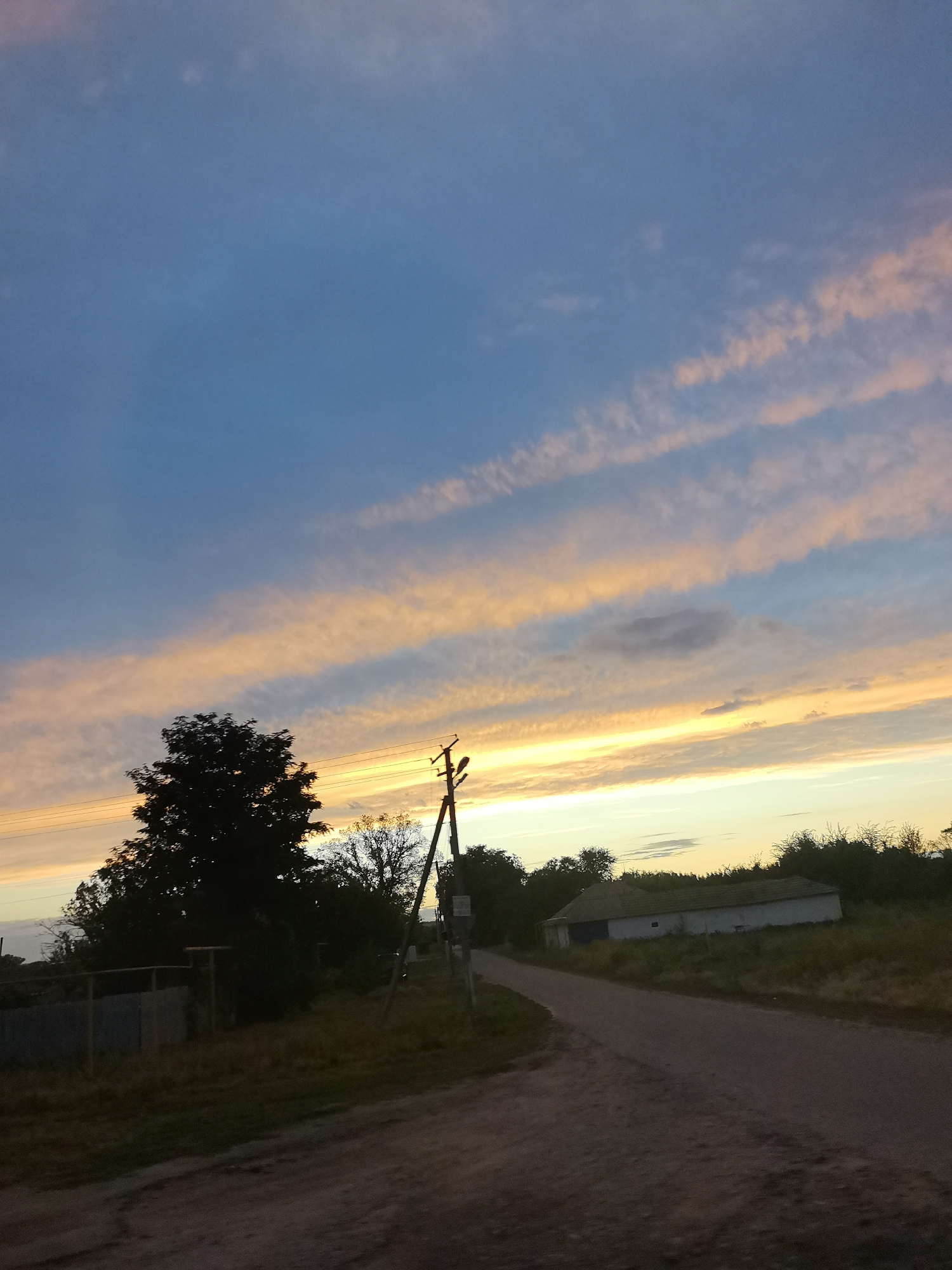 The image depicts a rural scene at dusk in Krasnoselovka, Russia. A dirt road curves to the right, leading past a dark silhouette of trees and a single-story building with a pitched roof. Power lines and a utility pole are visible on the right side of the road. The sky is a gradient of blue, transitioning to vibrant orange and pink hues from the setting sun, with wispy clouds stretching horizontally across the frame. The foreground is dominated by the unpaved road and a patch of dry grass.