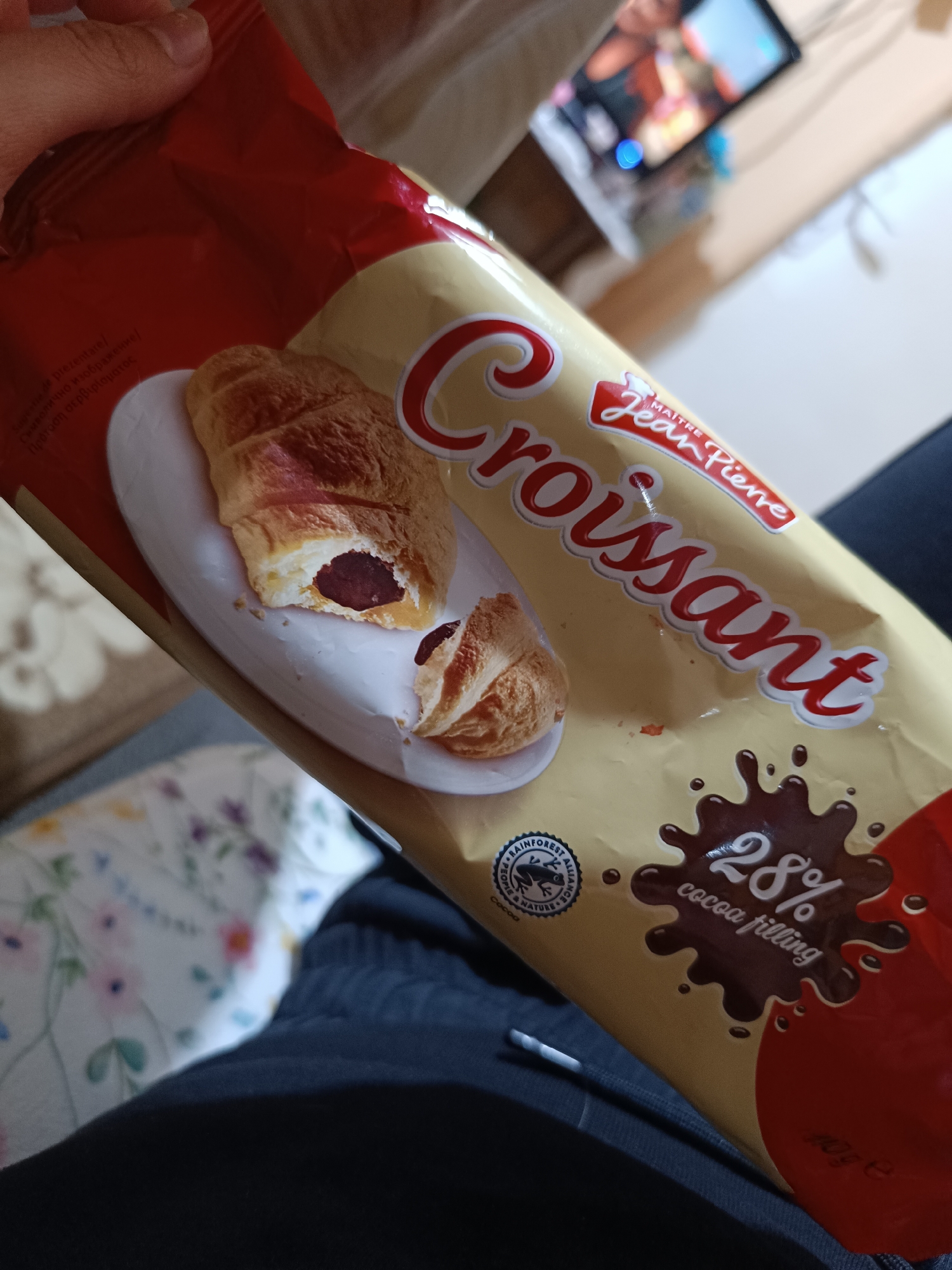The image shows a package of "Maitre Jean Pierre Croissant" with 28% cocoa filling. The croissant is depicted on the package with a portion cut away to reveal the cocoa filling. The packaging is primarily beige with red accents. The image suggests the croissant is being held by a person wearing dark clothing, and in the background, a television screen is visible. The image does not contain enough information to indicate the city of Pleven, Bulgaria.