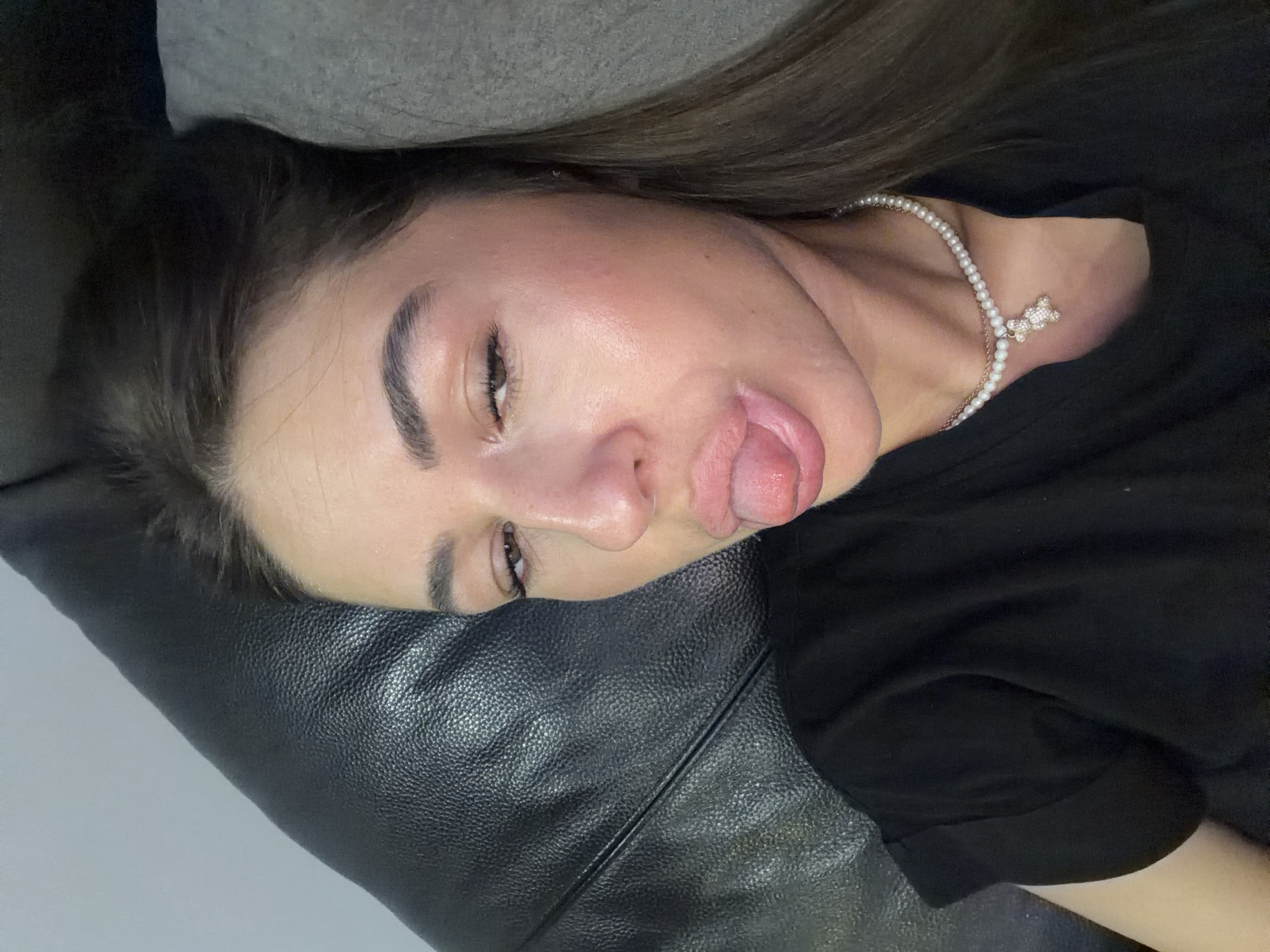A young woman is lying down, possibly on a couch or bed, with her head resting on a dark, textured surface, possibly leather, and a grey cushion behind her head. She is looking directly at the camera with her tongue sticking out. She is wearing a black t-shirt and a pearl necklace with a pendant shaped like a teddy bear adorned with rhinestones. The lighting suggests an indoor setting.