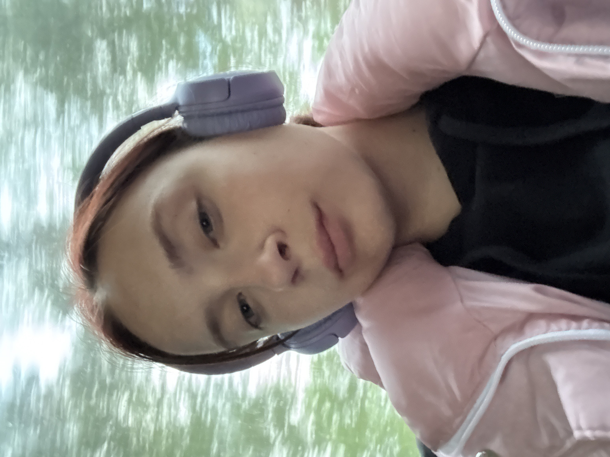 A young person with reddish-brown hair is wearing lavender over-ear headphones and a pink jacket. The background is blurred, possibly showing trees or an urban scene. The exact location in Lukintsy, Russia cannot be determined from the image alone.