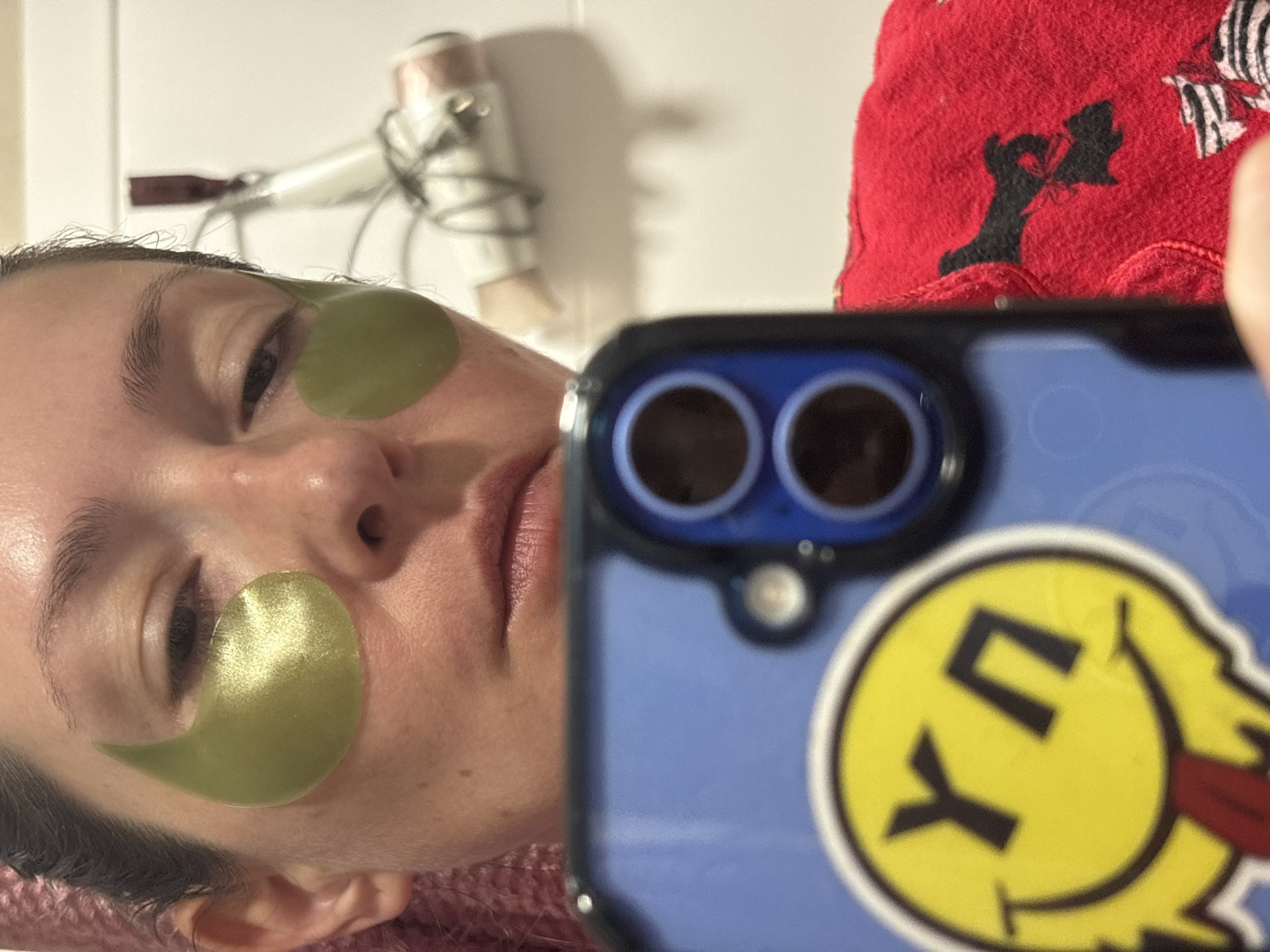 A selfie of a woman with green under-eye patches, indoors. A phone with a smiley face sticker is in the foreground, partially obscuring the view. In the background, there is a hairdryer mounted on a wall, and a red towel. The location is not discernible from the image.