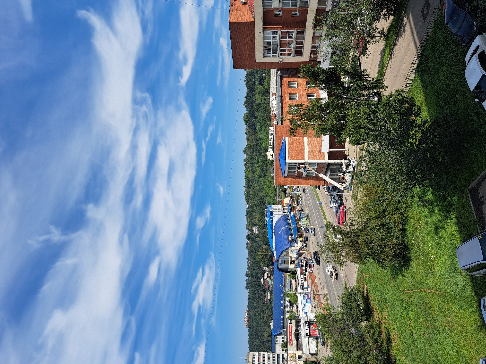 This is a vertical, eye-level, medium shot of a city street in Irkutsk, Russia. The image shows a multi-lane road with several cars driving and parked. The road is flanked by buildings of varying heights and architectural styles, some with commercial storefronts and others appearing to be residential. A large, modern building with a blue roof dominates the middle ground on the left side of the street, possibly a shopping center or indoor market. To the right, a multi-story brick building with large windows is visible. The background is filled with lush green trees, suggesting the presence of a park or forested area on a hillside. The sky is a clear blue with scattered white, wispy clouds. The overall impression is of a bustling urban environment on a sunny day.