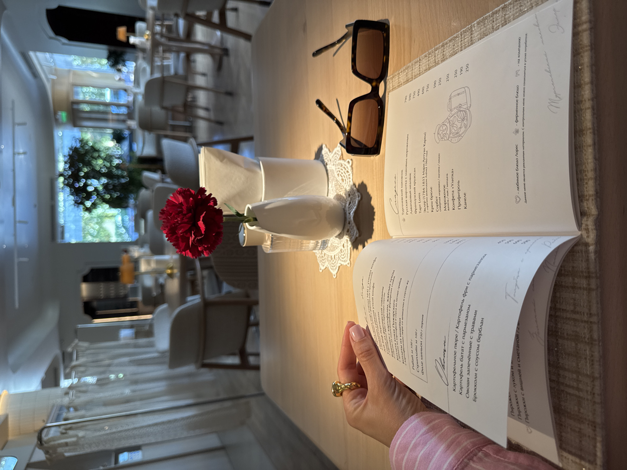 The image captures a first-person perspective at a restaurant table in Sochi, Russia. A person's hand, adorned with a gold ring, holds an open menu displaying Russian text. On the light wooden table, a pair of brown tortoiseshell sunglasses rests next to the menu. A small white vase holds a vibrant red carnation, positioned near a stack of white paper napkins contained in a simple holder, all resting on a delicate white lace doily.
The activity taking place is dining, with the individual perusing the menu to make a selection. The background reveals a bright, airy restaurant interior with natural light streaming through large windows, suggesting it is daytime. Outside, green foliage is visible, indicating an outdoor view. The setting appears modern and elegant.
Notable text from the menu includes various dishes and prices, primarily in Russian. Sections for desserts feature items like "Тургеневский маковник" (Turgenev poppy seed cake), "Лавандовый медовик" (Lavender honey cake), "Эклер" (Eclair), and "Мороженое" (Ice cream). Main courses include "Стейк из фермерской говядины" (Farm beef steak), "Котлеты из индейки" (Turkey cutlets), and "Рибай за 100 г" (Ribeye per 100g). Side dishes like "Картофельное пюре" (Mashed potatoes) and "Брокколи с соусом берблан" (Broccoli with beurre blanc sauce) are also listed. Phrases such as "любимое блюдо Лорес" (Lores' favorite dish) and "фирменное блюдо" (signature dish) are present, along with a disclaimer stating, "Данное меню является рекламным материалом. С контрольным меню можно ознакомиться в уголке потребителя." (This menu is advertising material. The control menu can be found in the consumer corner.). Handwritten signatures or notes are also visible on the menu pages.