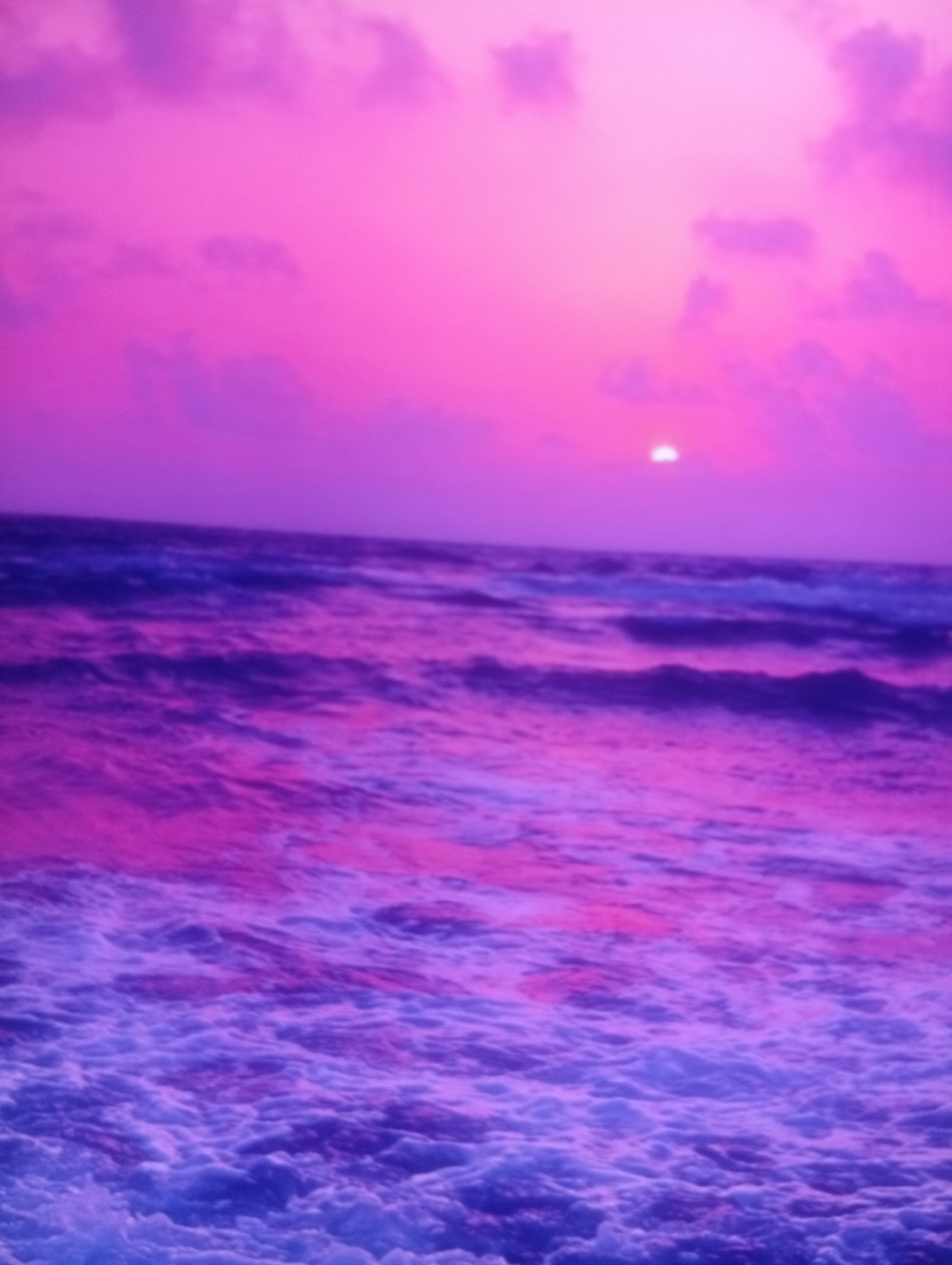 The image depicts a vibrant and ethereal sunset over a turbulent ocean. The sky is awash with shades of pink and purple, with soft, diffused clouds scattered across the horizon. The sun, a bright, glowing orb, is visible near the upper right corner, casting a warm, luminous glow. The ocean below is a dynamic expanse of deep blue and purple hues, with energetic waves crashing and frothing, creating a textured, almost painterly effect. The overall impression is one of dramatic natural beauty, with the intense colors and powerful movement of the water creating a sense of awe and wonder. There are no people or man-made objects visible in the frame, focusing solely on the raw elements of nature.