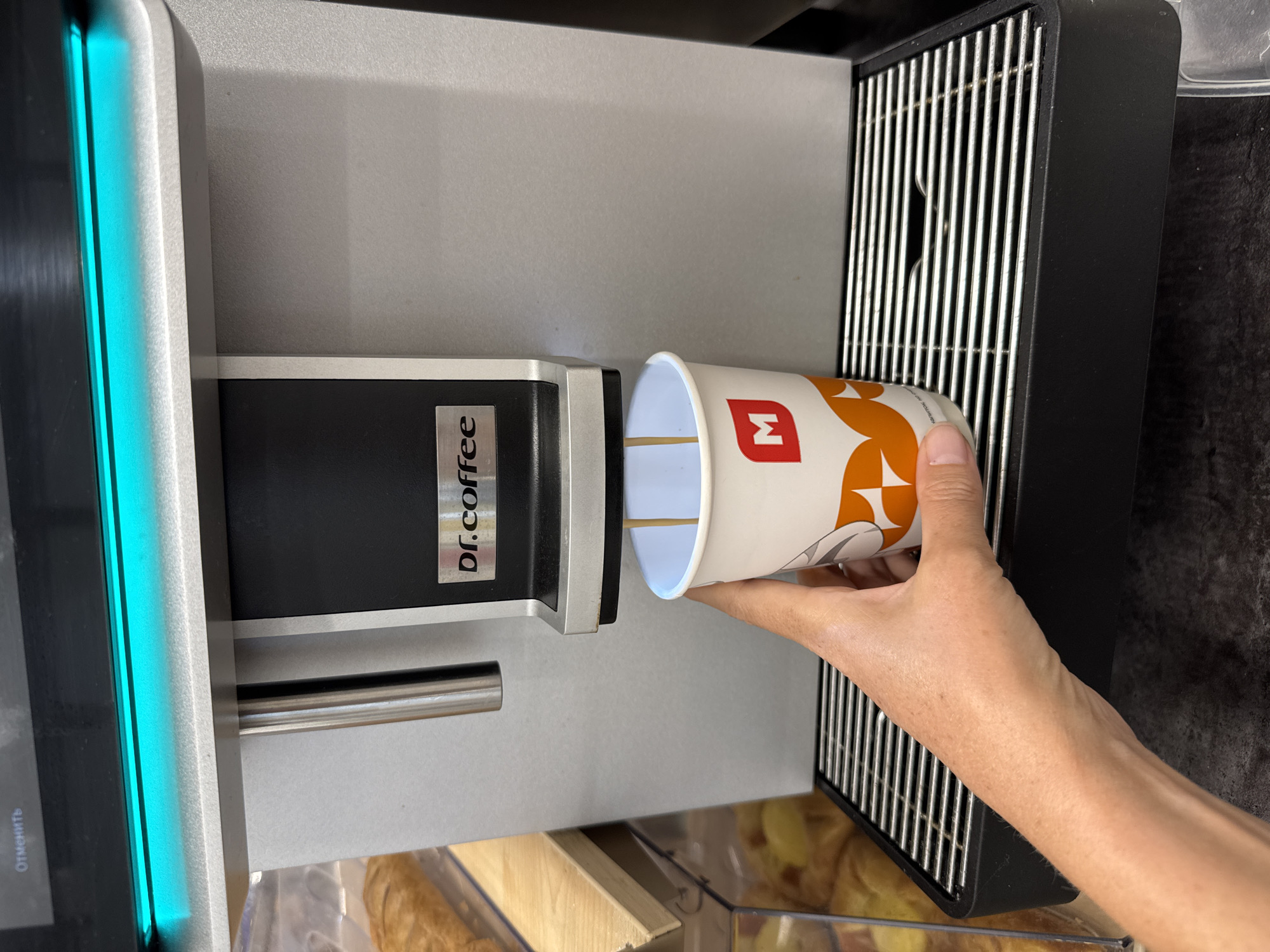 A person's hand is holding a paper coffee cup under a dispenser on a Dr. Coffee machine. The cup has an orange and white pattern and a red 'M' logo. The machine has a silver and black finish, with a bright blue light strip on the left side. The dispenser area has a metal grate. The background shows a display case with baked goods. The image is likely taken indoors, possibly in a cafe or convenience store. There is text on the machine that says "dr.coffee" and on the blue light strip which says "Отменить" (Cancel in Russian). The setting appears to be Kuivozi, Russia. The activity is the dispensing of coffee into a cup.