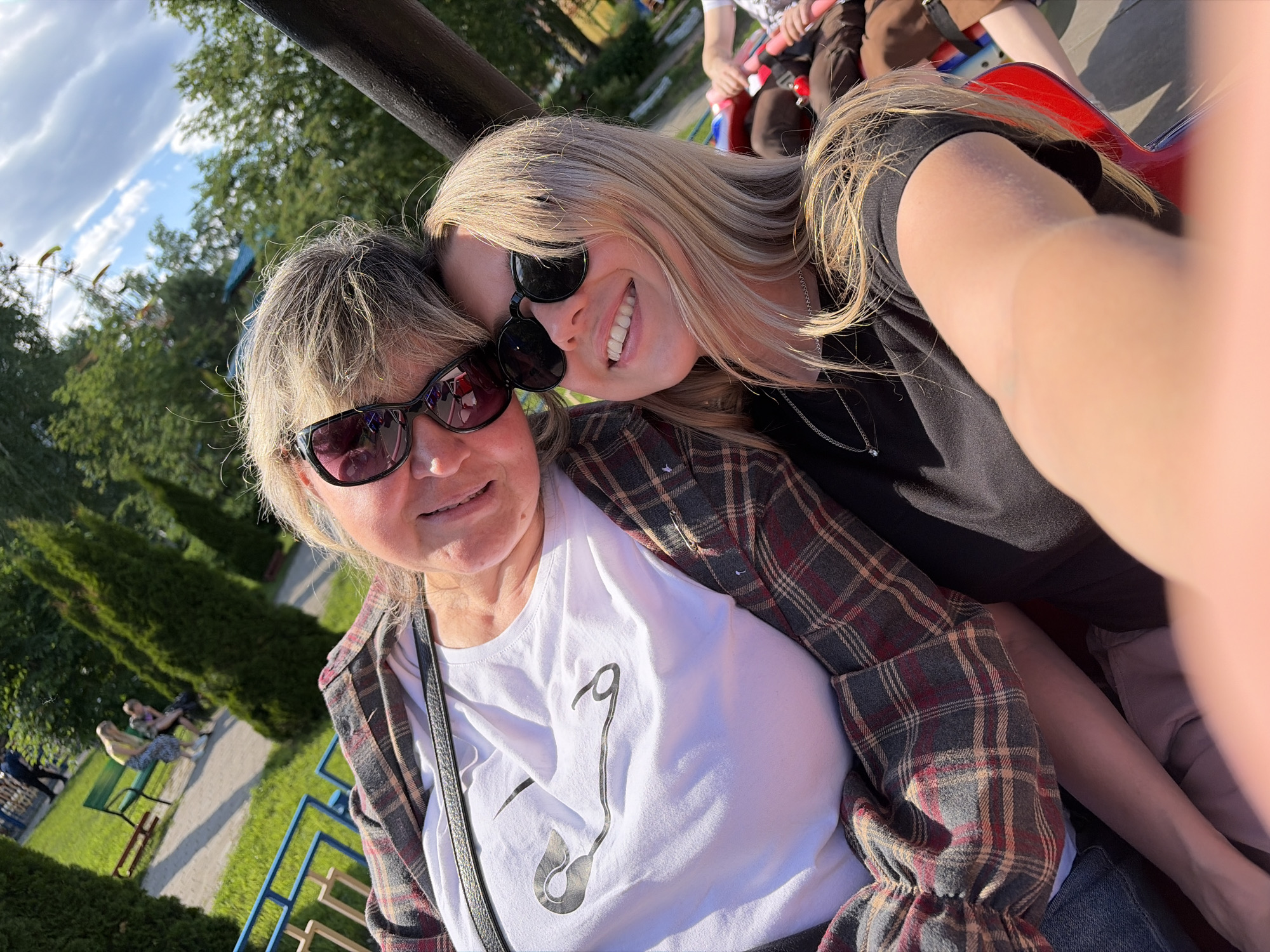 A selfie photograph captures two women in an outdoor amusement park or park environment, identified as being in Chaikovsky, Russia. The image is taken on a sunny day with a clear blue sky and scattered white clouds.
The woman in the foreground, positioned slightly to the left, has short, light blonde hair. She wears dark-framed sunglasses, a white short-sleeved t-shirt with a large black graphic depicting a safety pin, and an unbuttoned long-sleeved plaid shirt with a pattern of red, grey, and dark blue checks. A black bag strap crosses diagonally over her chest. She exhibits a subtle smile.
The woman positioned behind and to the right of the foreground woman, appearing younger, has long, straight blonde hair. She also wears dark-framed sunglasses and a black t-shirt or top with a delicate silver chain necklace. Her right arm is extended, visible on the far right of the frame, indicating she is holding the camera for the selfie. She displays a wide smile, revealing her teeth, and her head is angled towards the foreground woman's head, suggesting close interaction.
The background shows a green lawn with a paved pathway. Several people are visible in the mid-ground, some seated on a green park bench and others walking. Conical evergreen trees are present to the left. In the distance, various blurred amusement park structures, including yellow elements that could be part of a ride, are visible amidst green trees. A dark, thick metal bar runs diagonally across the top portion of the image. A red and blue structure is partially visible behind the younger woman, possibly part of a ride's seating or mechanism.