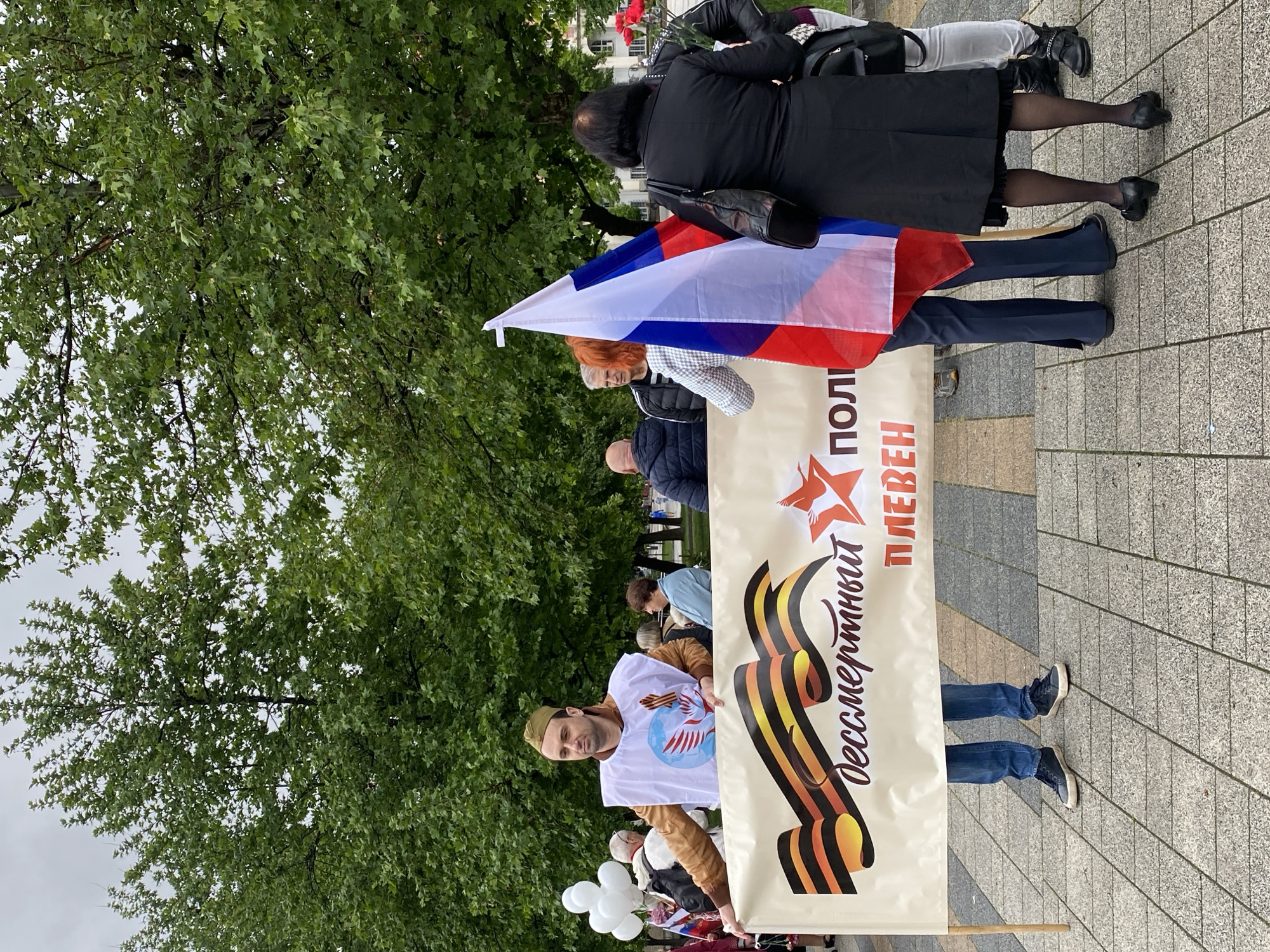 A street scene in Pleven, Bulgaria shows a group of people carrying a banner and the Russian flag. The banner has Cyrillic text and an image of a ribbon and a red star. One man wears a military cap and a t-shirt with an image on it. Several people are visible around the banner, and there are trees and pavement in the background.