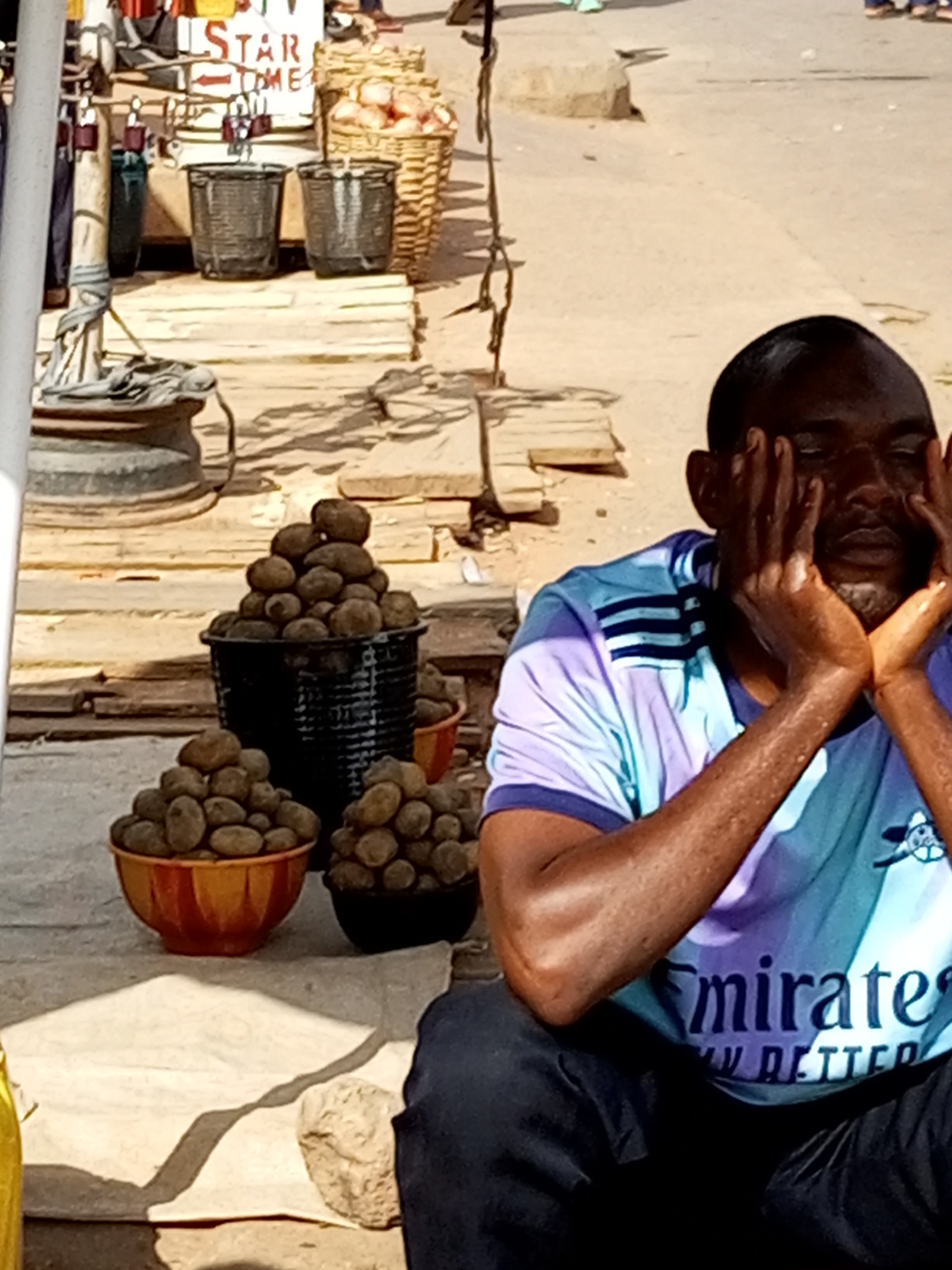 The image depicts a street scene in Tempure, Nigeria, focused on a man sitting on the ground with his hands covering his face. He is wearing a t-shirt with a "Fly Emirates" logo. In the foreground, there are three baskets of potatoes. Behind the man, a market stall is visible with various goods and baskets. The ground is dusty and appears to be a street or marketplace. The overall atmosphere suggests a busy outdoor environment.