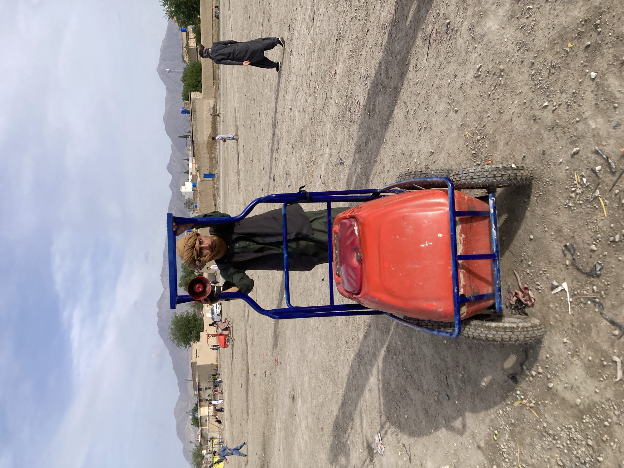 An outdoor scene in Quetta City Tehsil, Pakistan, depicts a large, dusty, unpaved open ground with scattered individuals. In the foreground, a male, appearing to be in his 20s or 30s, stands behind a blue, four-wheeled pushcart containing a horizontally mounted, red cylindrical tank. He wears a dark green shalwar kameez, a black vest, and a light brown turban, with a beard and mustache. He holds a red megaphone in his right hand, angled slightly upward and to his left, and looks directly at the camera.
In the midground, several individuals are visible across the unpaved area. To the right of the central figure, a male in a dark traditional outfit and black shoes walks away from the camera, wearing a dark head covering. To the far left, a child in a light blue tunic walks. Other adults and children are scattered across the background, some walking, others congregating near distant structures. A small, red, three-wheeled vehicle is visible in the mid-left background.
The background consists of a row of single-story, light-colored buildings with flat roofs, some featuring blue doors or accents. Beyond these structures, a range of rugged, barren mountains extends across the horizon under a partly cloudy, light blue sky. A utility pole is faintly visible against the mountain range on the right side. The ground is light brown, dry, and littered with small stones and debris, suggesting an arid environment.