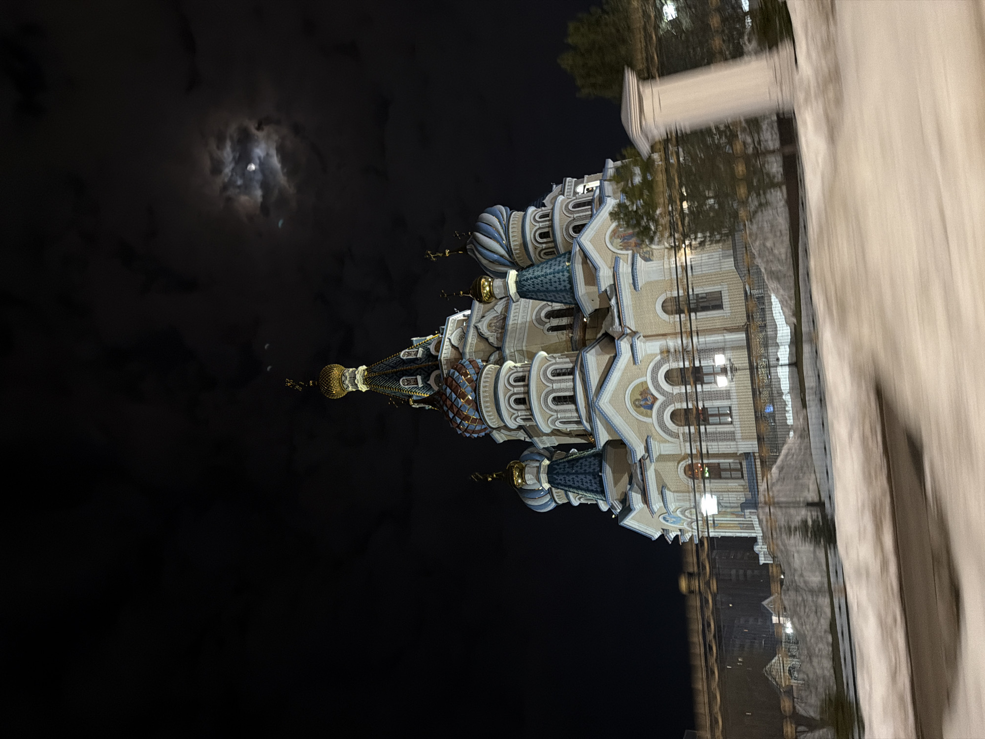 The image shows St. Michael's Cathedral in Izhevsk, Russia, at night, under a cloudy sky with the moon visible. The cathedral is brightly lit, showcasing its architectural details, including the onion domes. Part of a light-colored wall is visible in the right foreground, blurring slightly, suggesting the photograph might have been taken from a moving vehicle or with a handheld camera. The rest of the background is dark, with some lights visible.