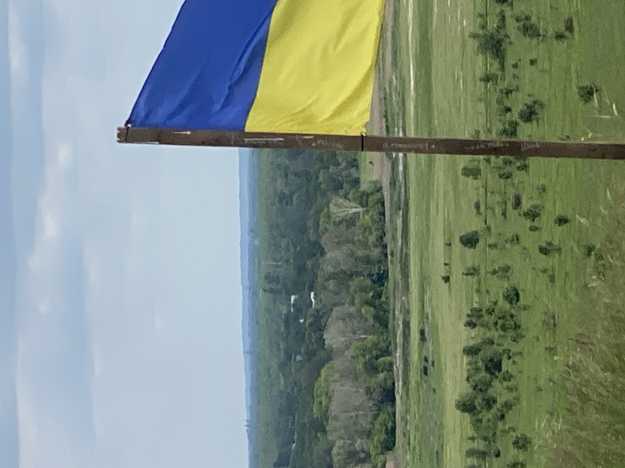 The image shows a Ukrainian flag waving from a pole. The sky is overcast. In the background, a landscape of green fields and wooded areas stretch towards the horizon. Buildings are visible amidst the trees. The location is Ustyvytsia, Ukraine.