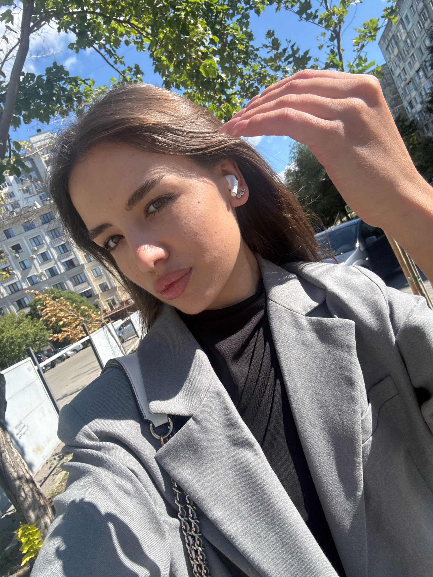In Chișinău, Moldova, a young woman with dark brown hair and brown eyes is captured in a close-up selfie outdoors. She is wearing a grey blazer over a black top and white wireless earbuds in her left ear. Her right hand is raised, gently touching her hair. The background features buildings, trees, and a bright blue sky. The lighting suggests it is daytime.