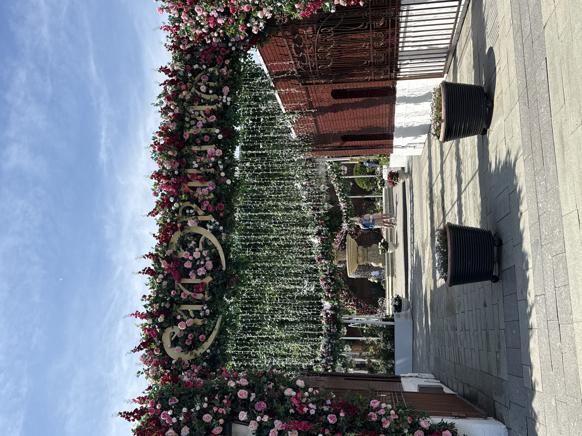 This image captures an outdoor, elaborately decorated entrance or walkway under a clear sky in Moscow, Russia, at a venue identifiable as "Сан-РЕМO" (San-Remo).
In the immediate foreground, a walkway composed of light grey rectangular paving stones extends towards the background. Two dark-colored, ribbed cylindrical planters, each equipped with small wheels and containing green plants, are placed symmetrically on the walkway. Shadows are cast onto the pavement, indicating strong overhead sunlight.
A prominent archway, heavily adorned with a dense array of pink, red, and white roses, interspersed with red campanulate flowers and green foliage, spans the width of the path. Centered within the top section of this floral archway is a large, golden-colored inscription in Cyrillic, reading "САН-РЕМO". Hanging vertically below the archway are numerous strands of white lights intertwined with artificial green vines, creating a hanging garden effect.
To the left of the archway, a section of a dark red brick building is partially obscured by cascading floral decorations. A dark metal gate or railing with ornate scrollwork is visible alongside this building.
To the right, a dark red brick building features two recessed arched openings. A dark metal railing runs along its base, and similar floral decorations extend downwards from the archway along this side.
Beyond the archway, in the midground, a multi-tiered light-colored stone fountain with water actively flowing is positioned slightly to the right of center. Further back, additional white structures, possibly decorative arches or pergolas, also adorned with flowers, are visible. Green trees and shrubs form the background.
Two individuals are standing near the fountain on the right side: a male in a blue collared shirt and beige shorts, and a female in a light pink top and light-colored shorts. They are positioned facing towards the fountain area. A third person in lighter attire is partially visible further back near the fountain.
The upper portion of the image reveals a clear, light blue sky with scattered wispy white clouds. A faint, small crescent object, consistent with a daytime moon, is discernible in the upper central area of the sky. The entire scene is brightly illuminated by natural daylight.