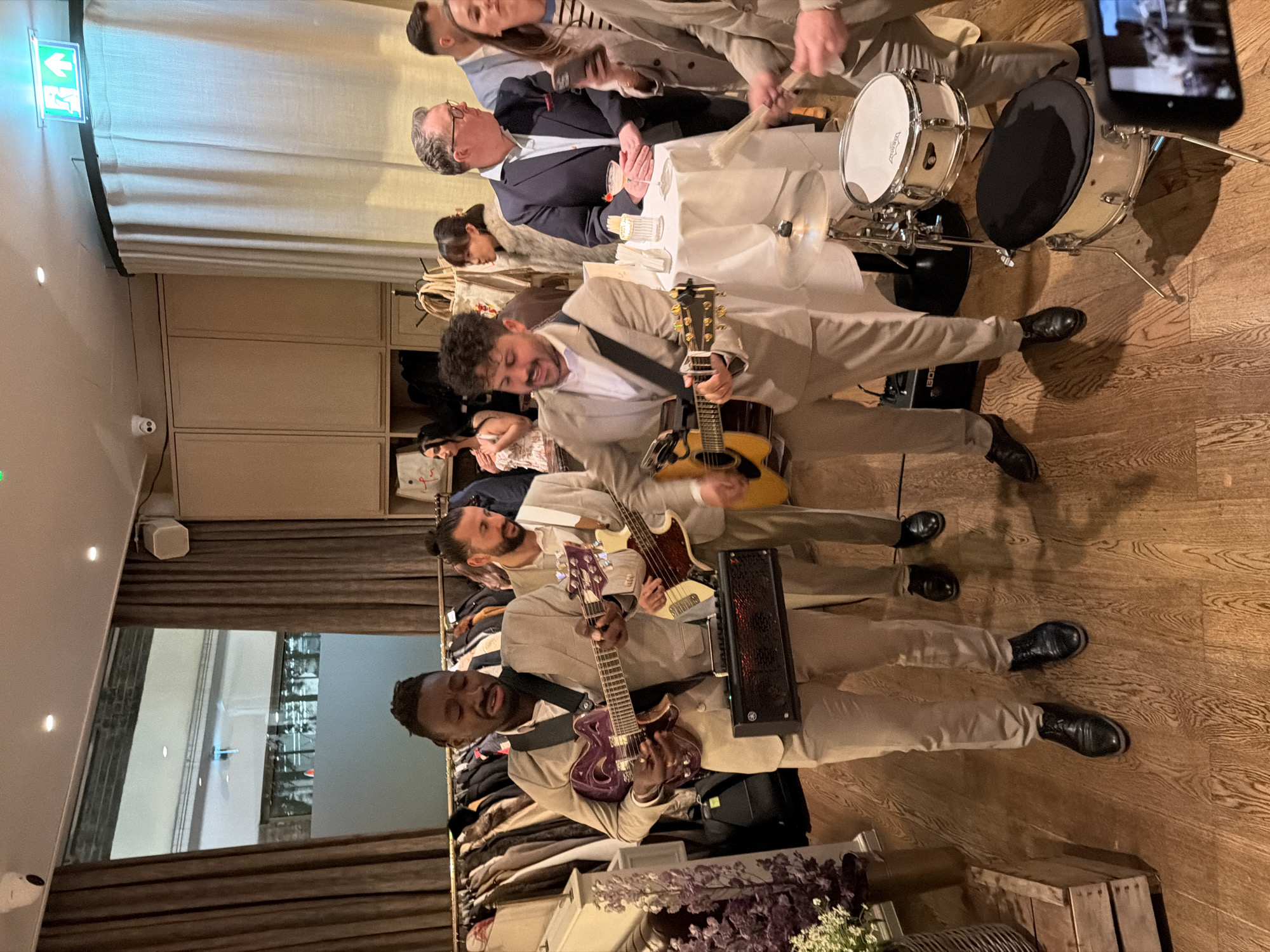 The image portrays an indoor event taking place in a retail or hospitality venue in London, United Kingdom. In the foreground, three male musicians, each wearing a light-colored suit, are actively performing. The male on the left plays a purple semi-hollow body electric guitar and stands near a rectangular black amplifier with an illuminated red grille. A second male, positioned behind the first, plays a light-colored bass guitar. The third male, on the right, plays an acoustic guitar. A partial drum kit, consisting of a white snare drum and a white floor tom, is visible in the lower right, with drum brushes resting on the snare.
In the midground and background, multiple individuals are present, observing the performance or standing near small, round tables draped with white tablecloths. One table holds a cocktail garnished with a cherry and a small decorative item. To the left, a clothing rack displays numerous garments, including jackets and coats. The venue features light wooden flooring, light beige wall panels, and matching drapes. Overhead, multiple recessed lights illuminate the space, and a green illuminated exit sign is visible. A white security camera is mounted on the ceiling. A large window with a frosted lower section is visible in the background, revealing an obscured architectural detail.