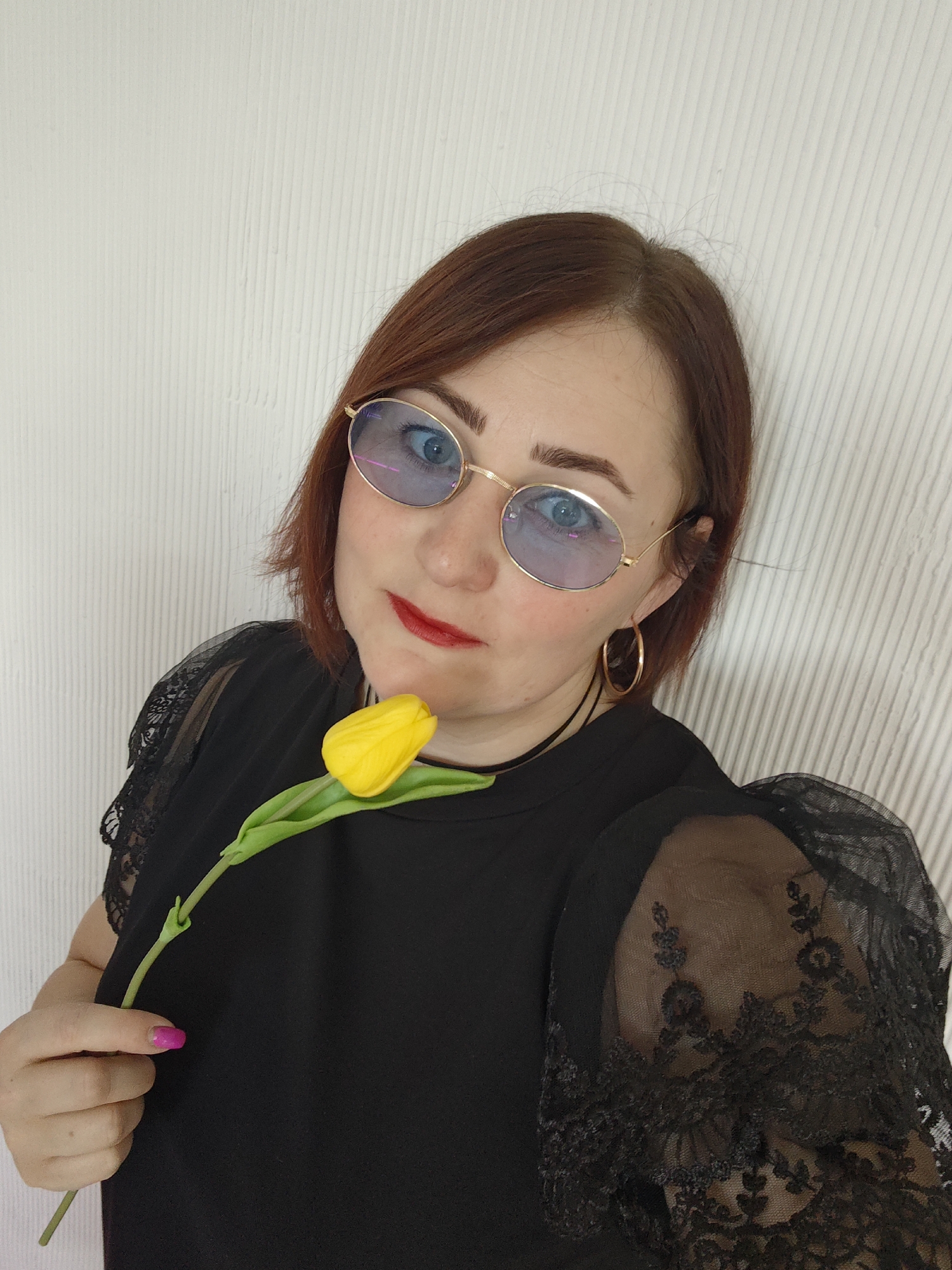 An indoor, mid-shot portrait captures an adult female situated in Chaikovsky, Russia, directly facing the camera. She has medium-length reddish-brown hair and wears oval-shaped glasses with gold frames and blue-tinted lenses, through which her left eye is visible. Her lips are adorned with red lipstick. She is dressed in a black top featuring sheer, lace-patterned sleeves and wears a black choker necklace along with a gold-colored hoop earring on her right ear. Her visible left hand, with bright pink fingernail polish, holds a single yellow tulip with a green stem, positioned near her chin. The background is a light-colored wall displaying a consistent vertical ribbed texture.
